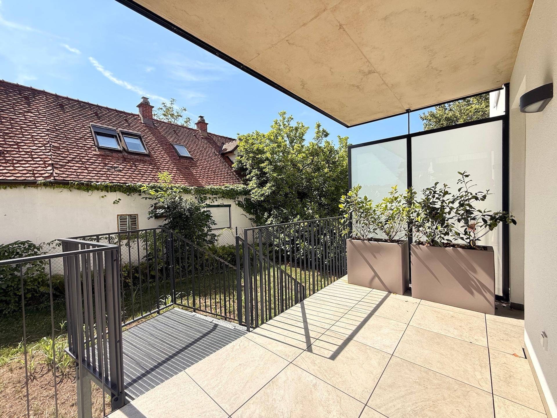 Helle Terrasse mit Fliesenboden, Pflanzenkübeln und Blick auf umliegende Gebäude und Grünflächen.