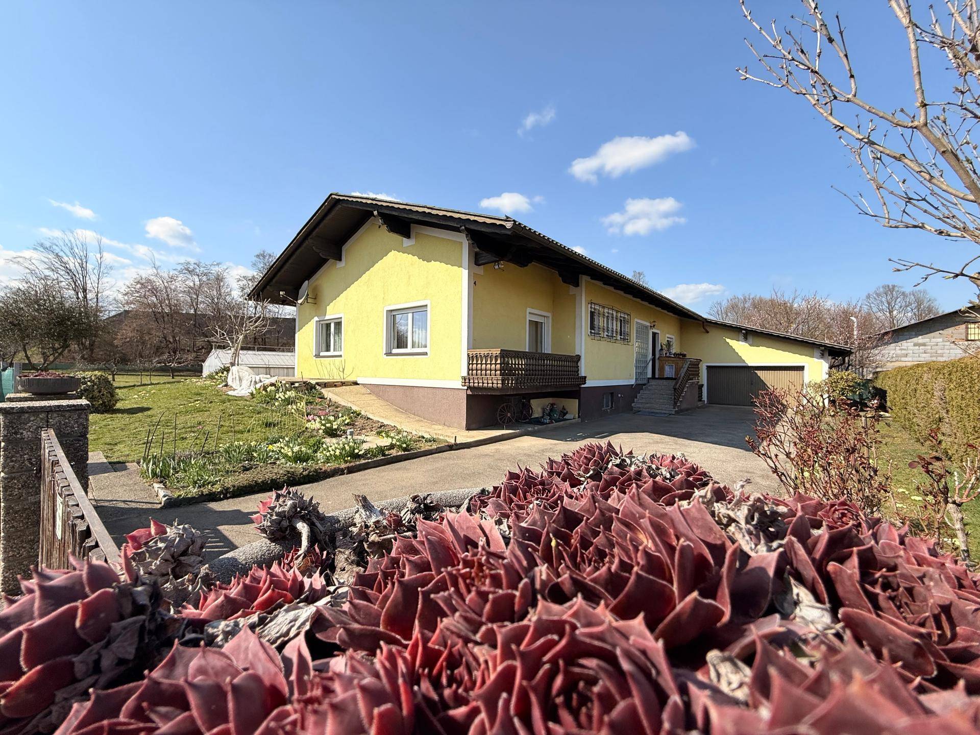 Gelbes Einfamilienhaus mit Satteldach, umgeben von einem Garten und einer Einfahrt an einem sonnigen Tag.