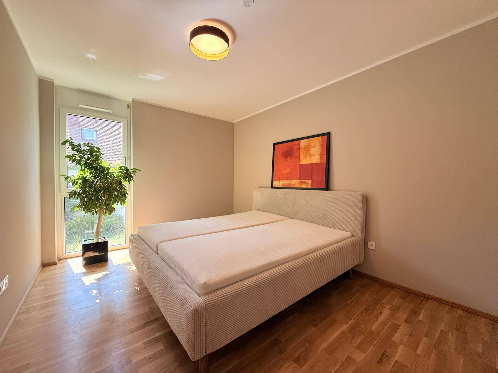 Helles Schlafzimmer mit Doppelbett, Parkettboden und Fenster mit Blick ins Grüne.