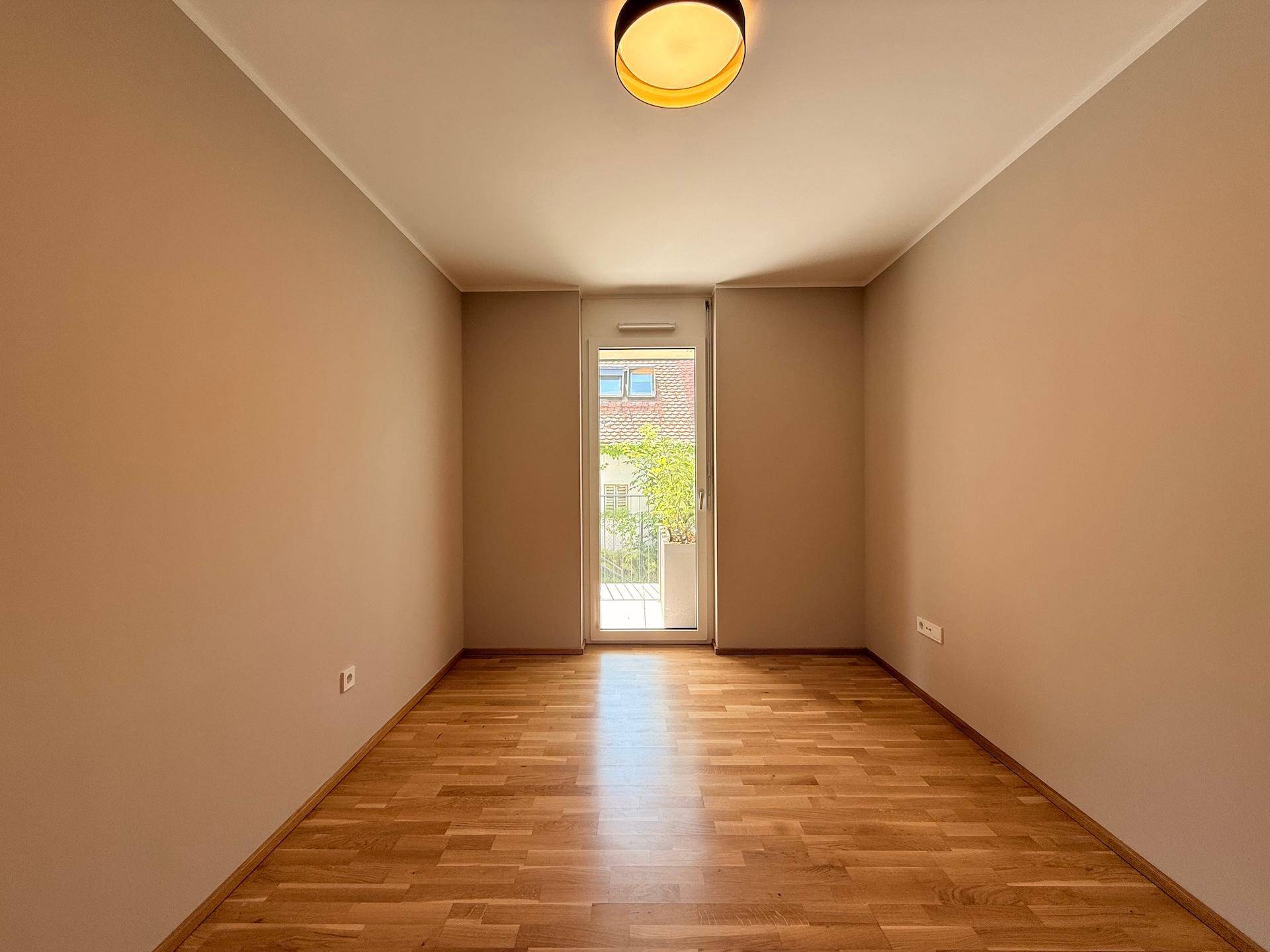 Leeres Zimmer mit hellem Holzparkettboden und Zugang zu einem kleinen Balkon.