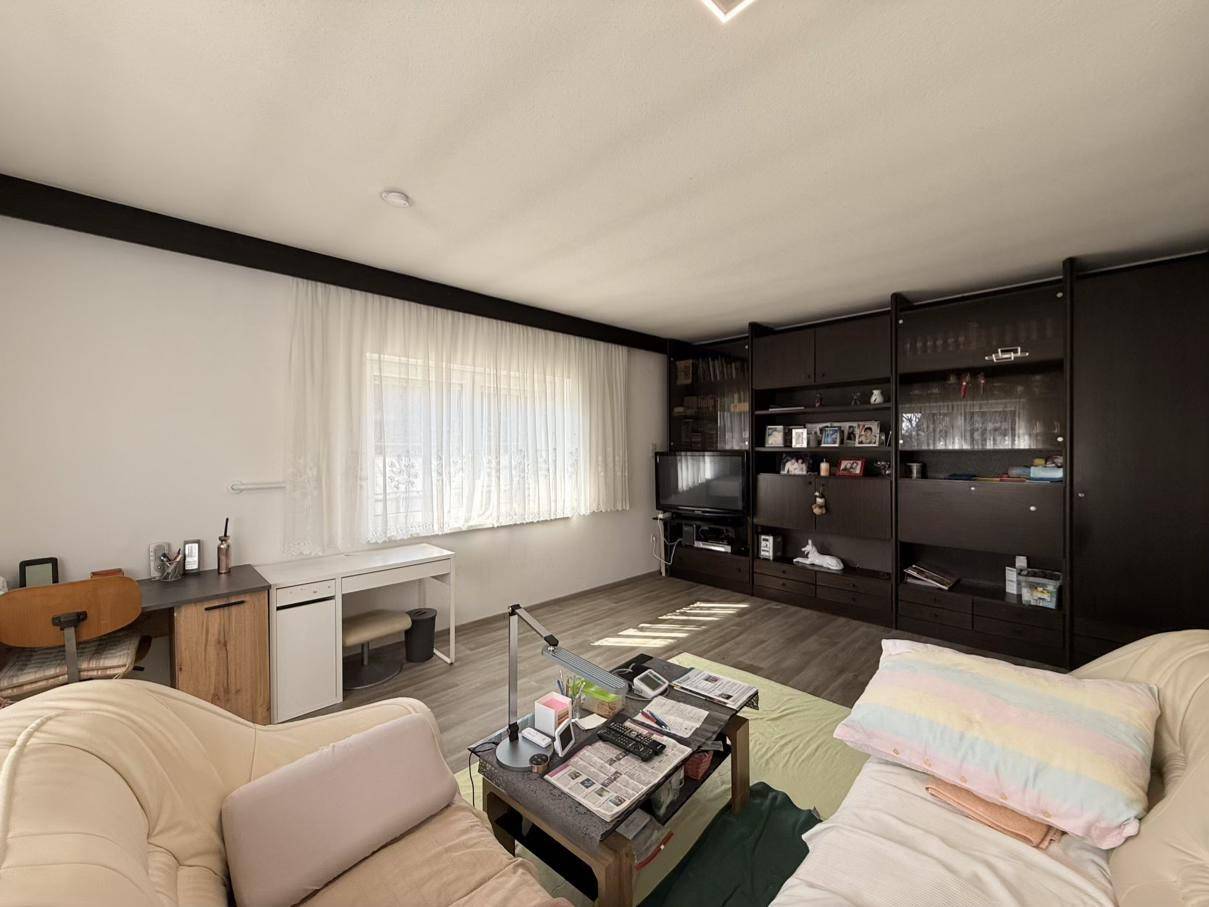Wohnzimmer mit beigem Sofa, Schrankwand und einem Arbeitsbereich, hell durch Tageslicht.