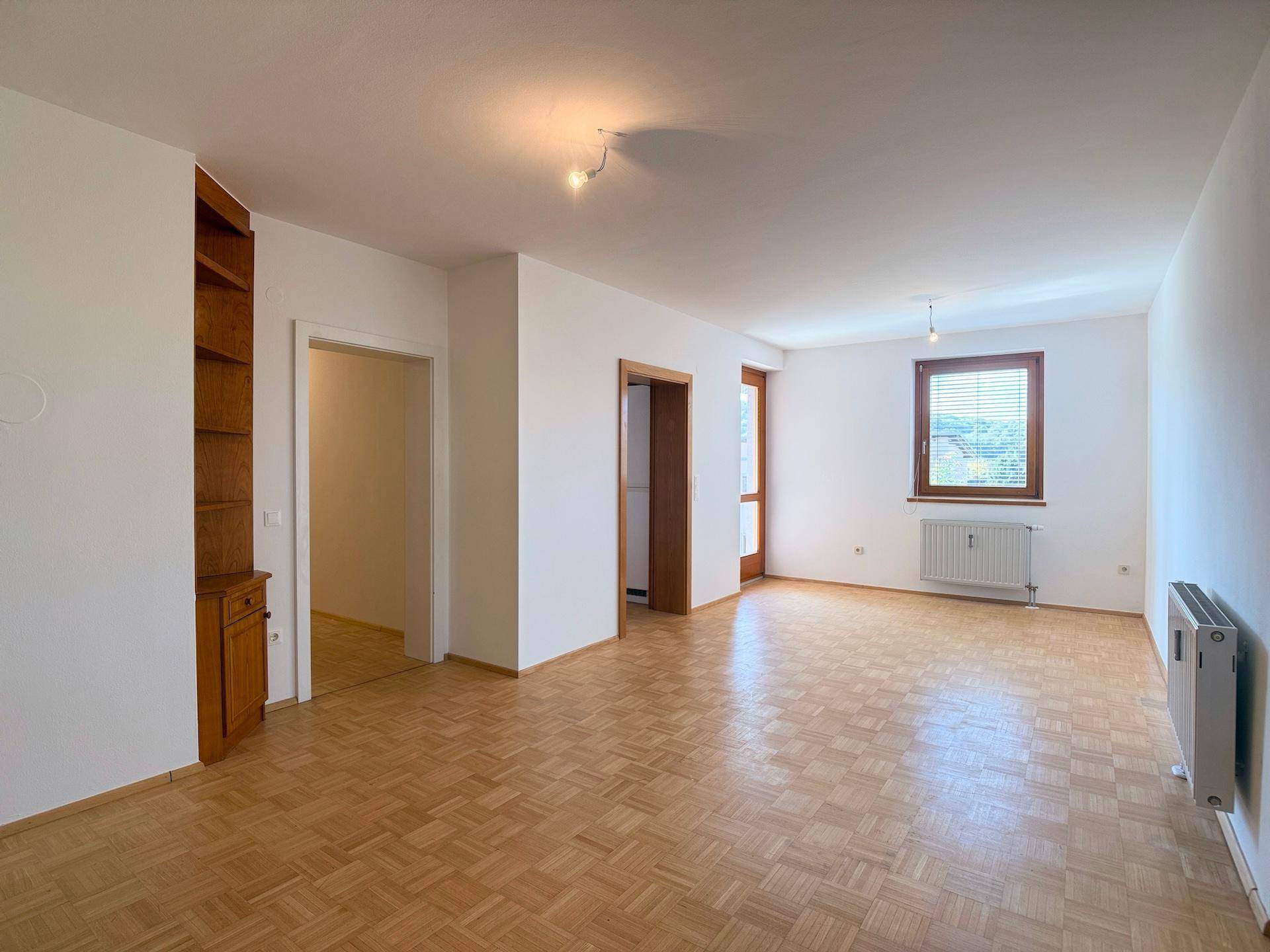 Geräumiges Wohnzimmer mit hellem Parkettboden und einem Fenster, das viel Tageslicht hereinlässt.
