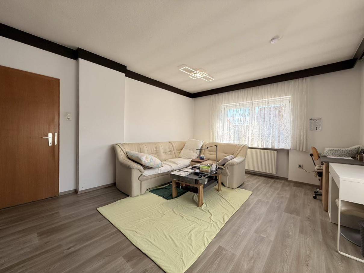 Wohnzimmer mit beigem Sofa und Laminatboden, hell durch ein großes Fenster.