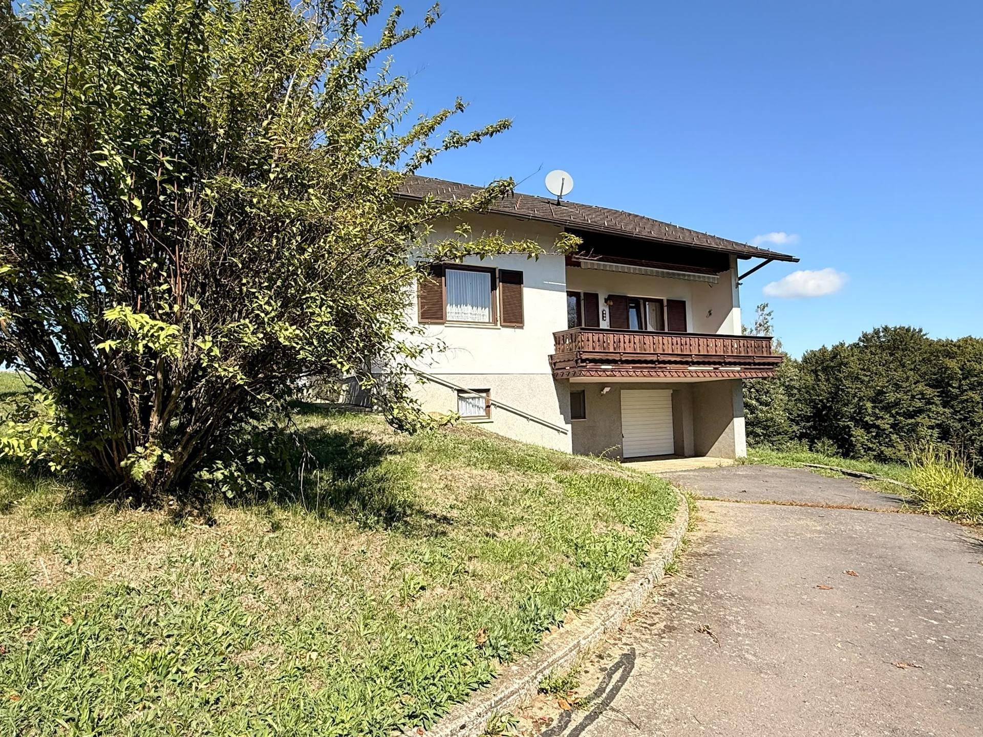 Ansicht des Hauses von der Rückseite mit Balkon, Garage und Zufahrt, umgeben von Bäumen.