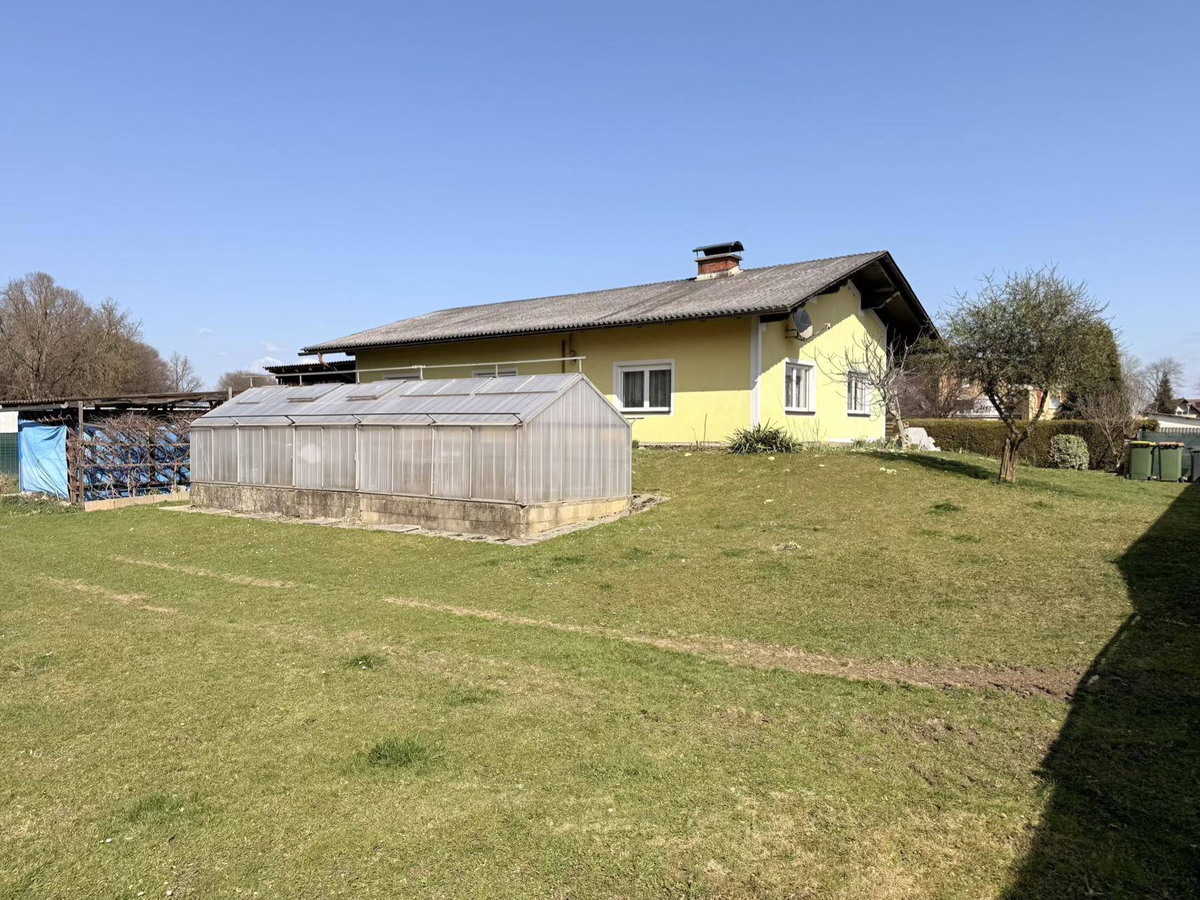 Gartenbereich mit einem Gewächshaus und dem gelben Haus im Hintergrund, umgeben von einer Rasenfläche.