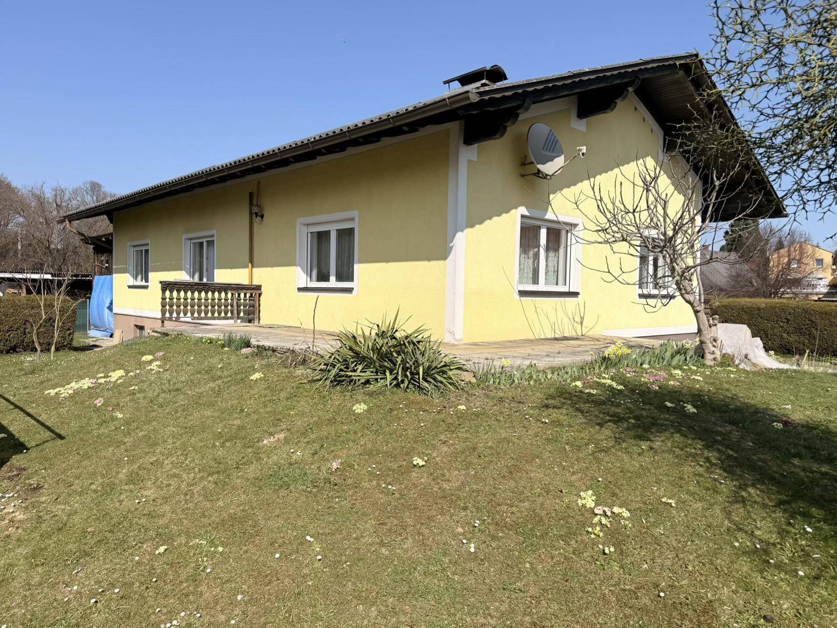 Gelbes Haus mit gepflasterter Fläche neben dem Gebäude und einem grünen Rasenbereich an einem sonnigen Tag.