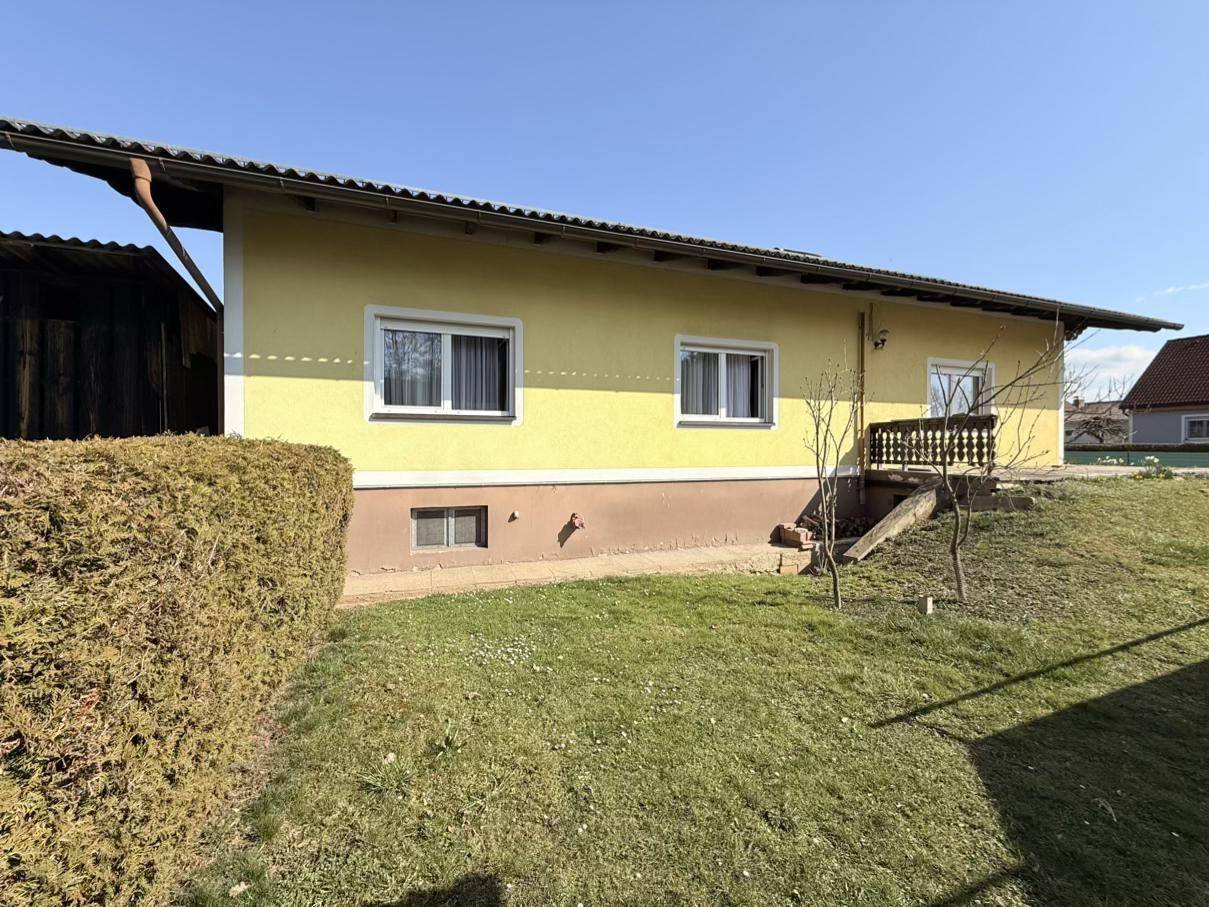 Seitenansicht des gelben Hauses mit einer gepflegten Hecke und einem grünen Rasenbereich im Sonnenschein.