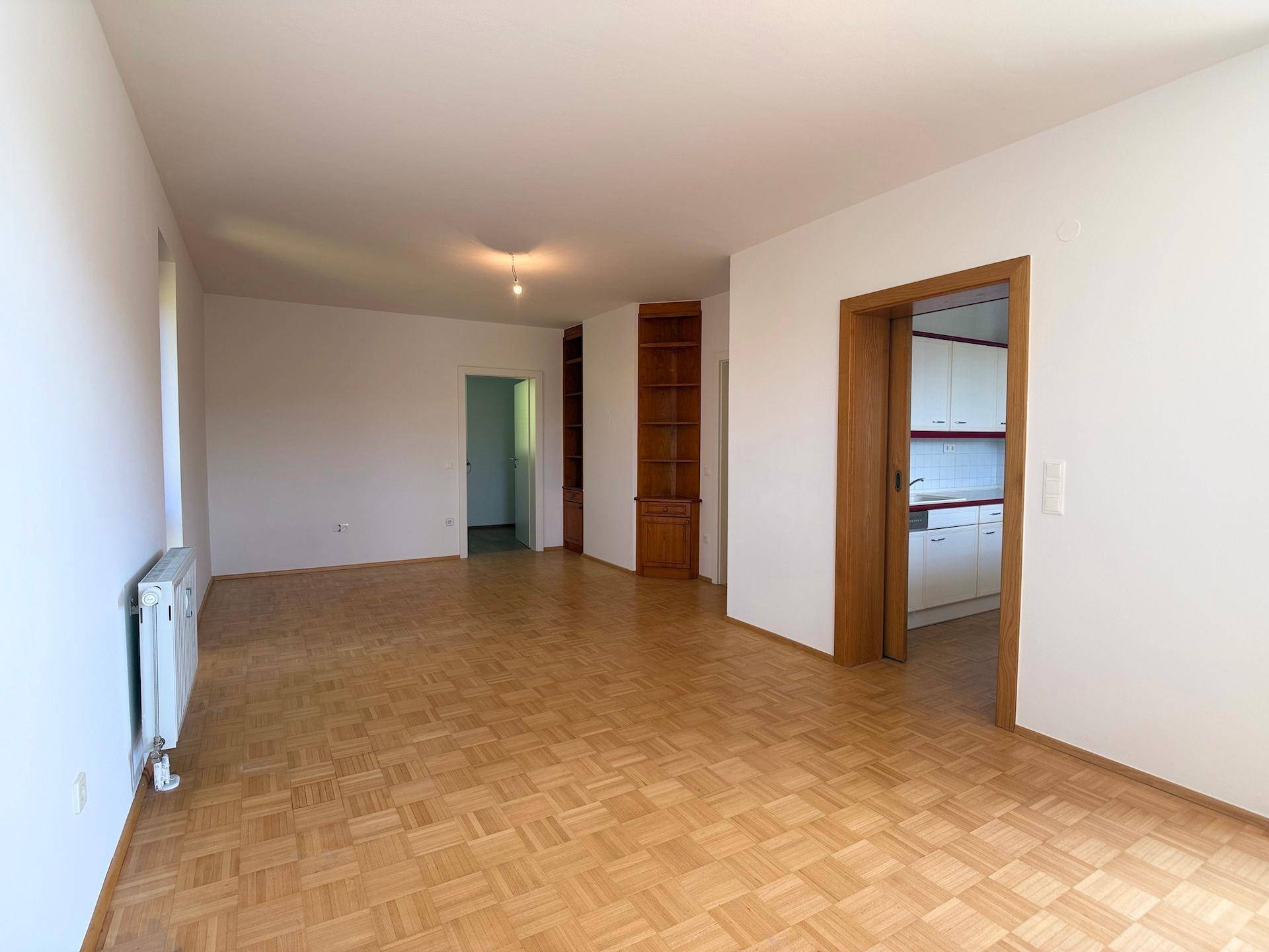 Helles Wohnzimmer mit Parkettboden und Zugang zu weiteren Räumen, inklusive der Küche.