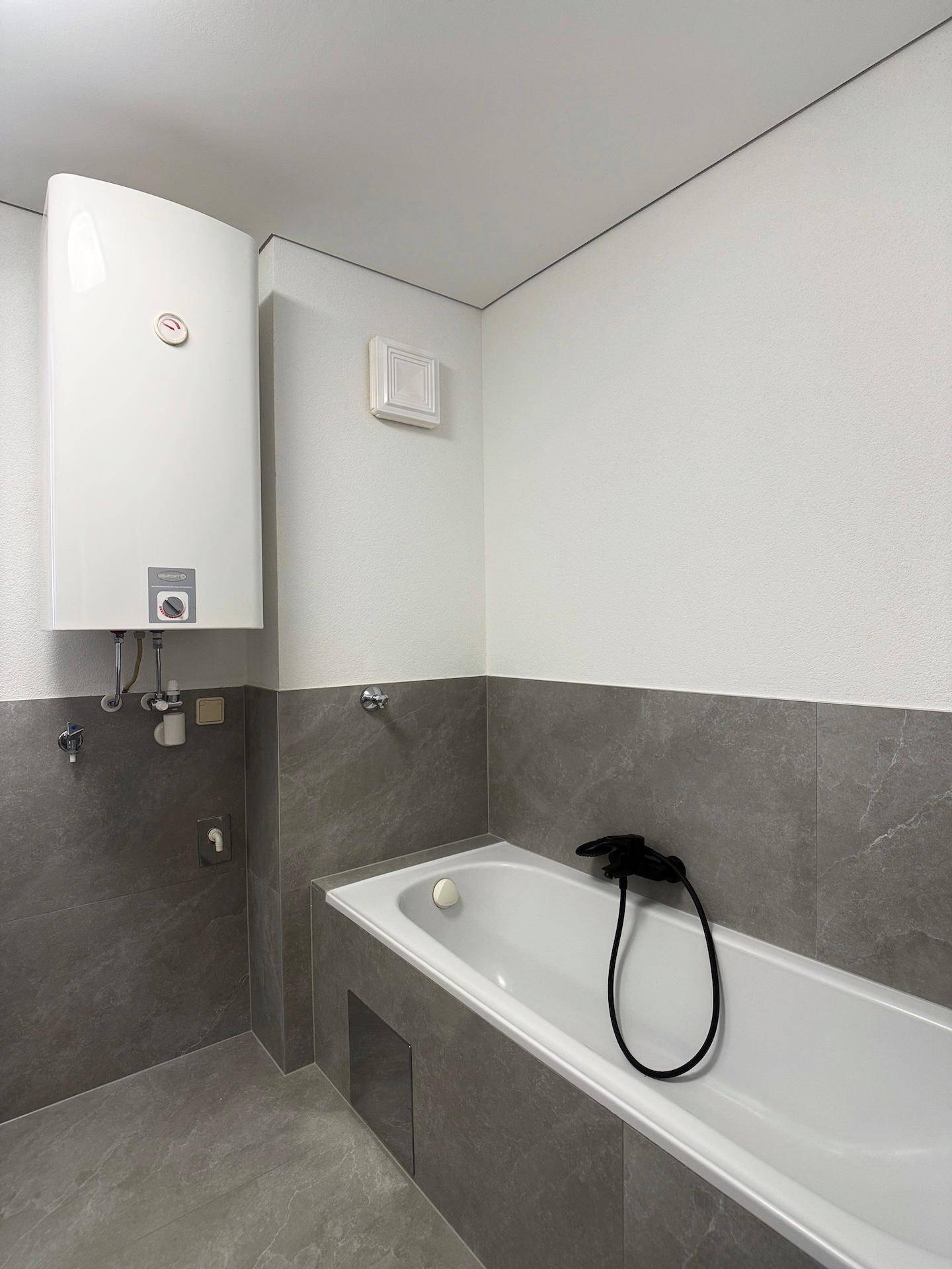 Modernes Badezimmer mit Badewanne, grauen Fliesen und einem Warmwasserboiler an der Wand.