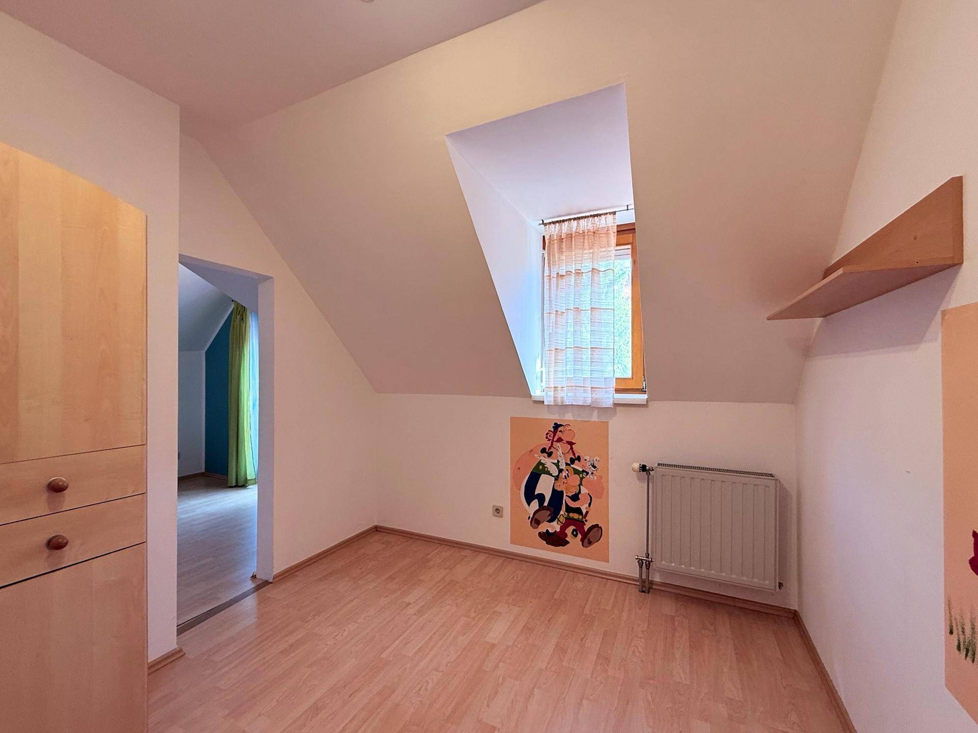 Helles Zimmer mit Dachschräge, Laminatboden und Fenster mit Vorhängen.
