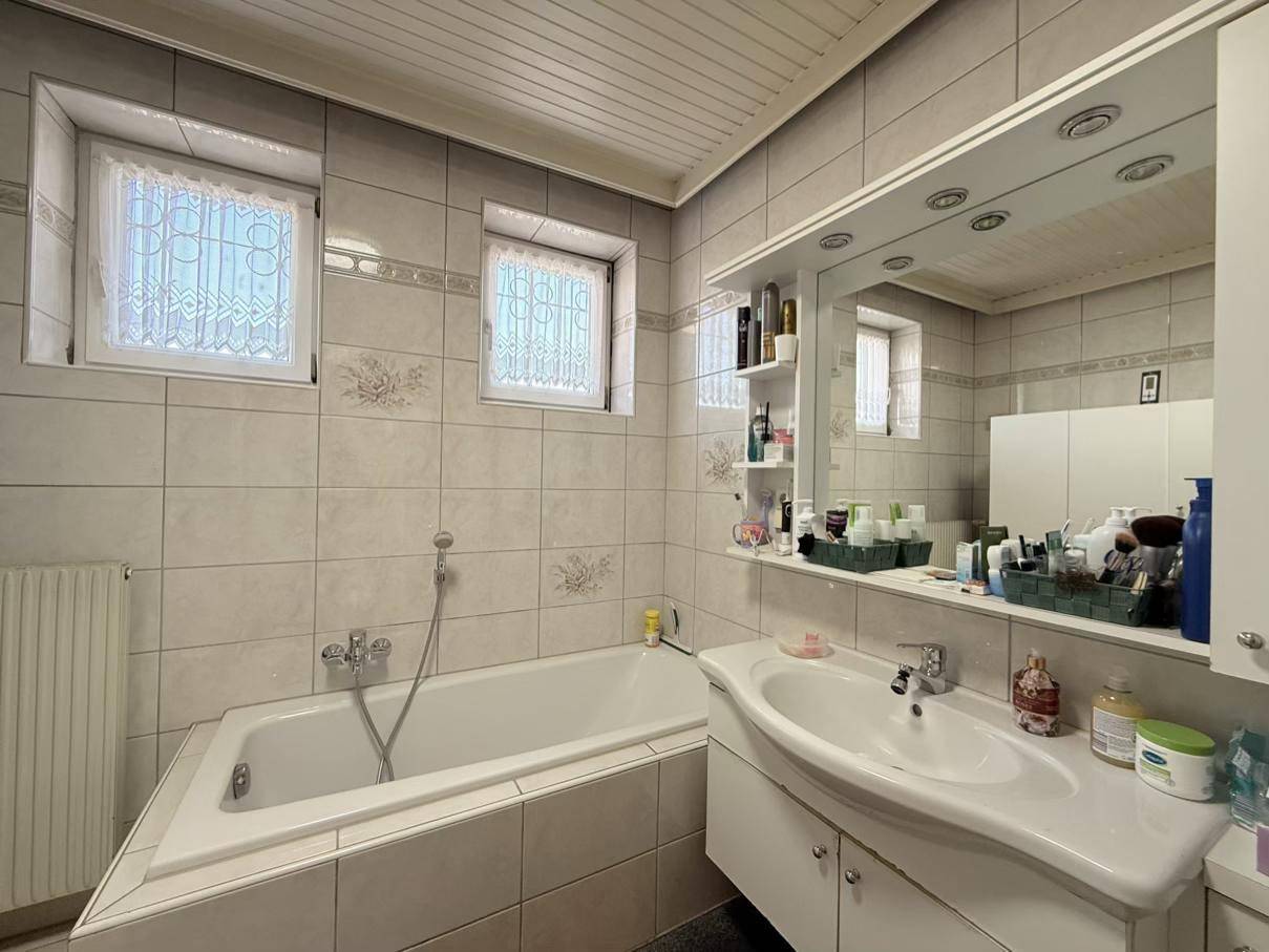 Gefliestes Badezimmer mit Badewanne, Waschbecken und Spiegelschrank, beleuchtet durch zwei Fenster.