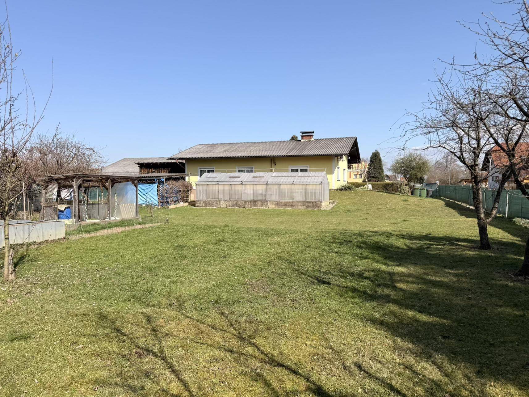 Weitläufiger Garten mit Rasenfläche, Gewächshaus und dem gelben Haus im Hintergrund an einem sonnigen Tag.