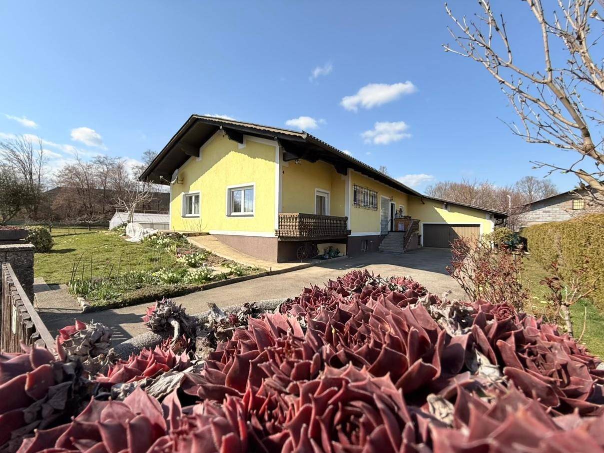 Gelbes Einfamilienhaus mit Satteldach, umgeben von einem Garten und einer Einfahrt an einem sonnigen Tag.
