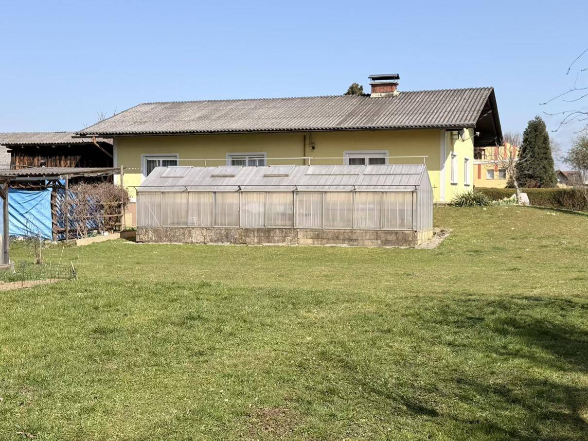 Gartenbereich mit einem Gewächshaus und dem gelben Haus im Hintergrund, umgeben von einer Rasenfläche.