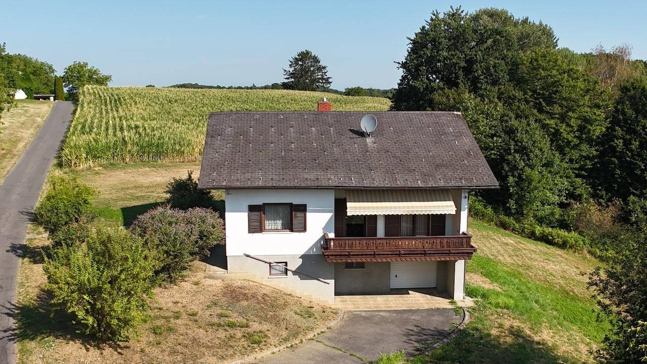 Frontale Luftaufnahme des Hauses mit Balkon und Garageneinfahrt, umgeben von grünen Hügeln.