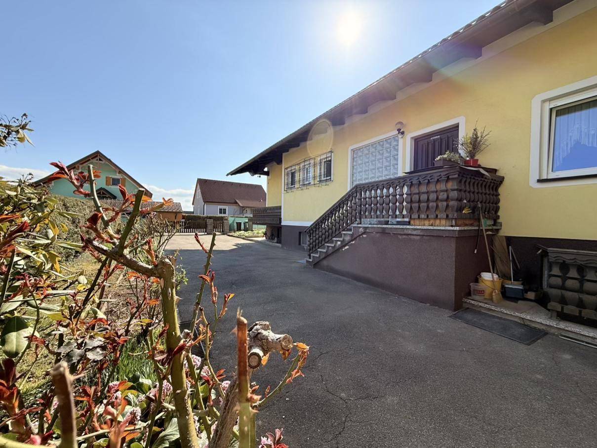 Eingangsbereich des gelben Hauses mit Holzgeländer und dekorativen Pflanzen im Vordergrund bei Sonnenschein.