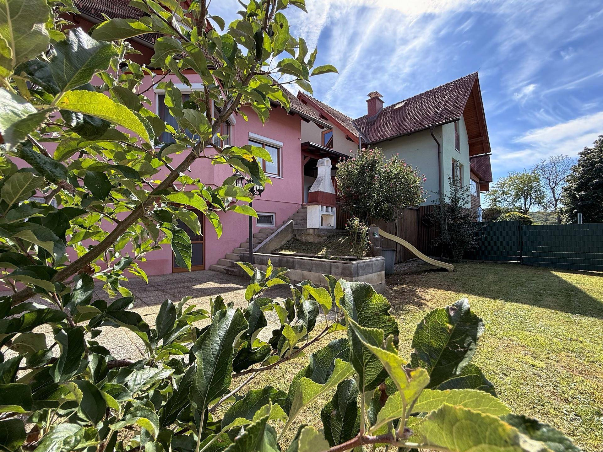 Blick auf das rosa Haus vom Garten aus, umgeben von grünen Bäumen und Sträuchern.