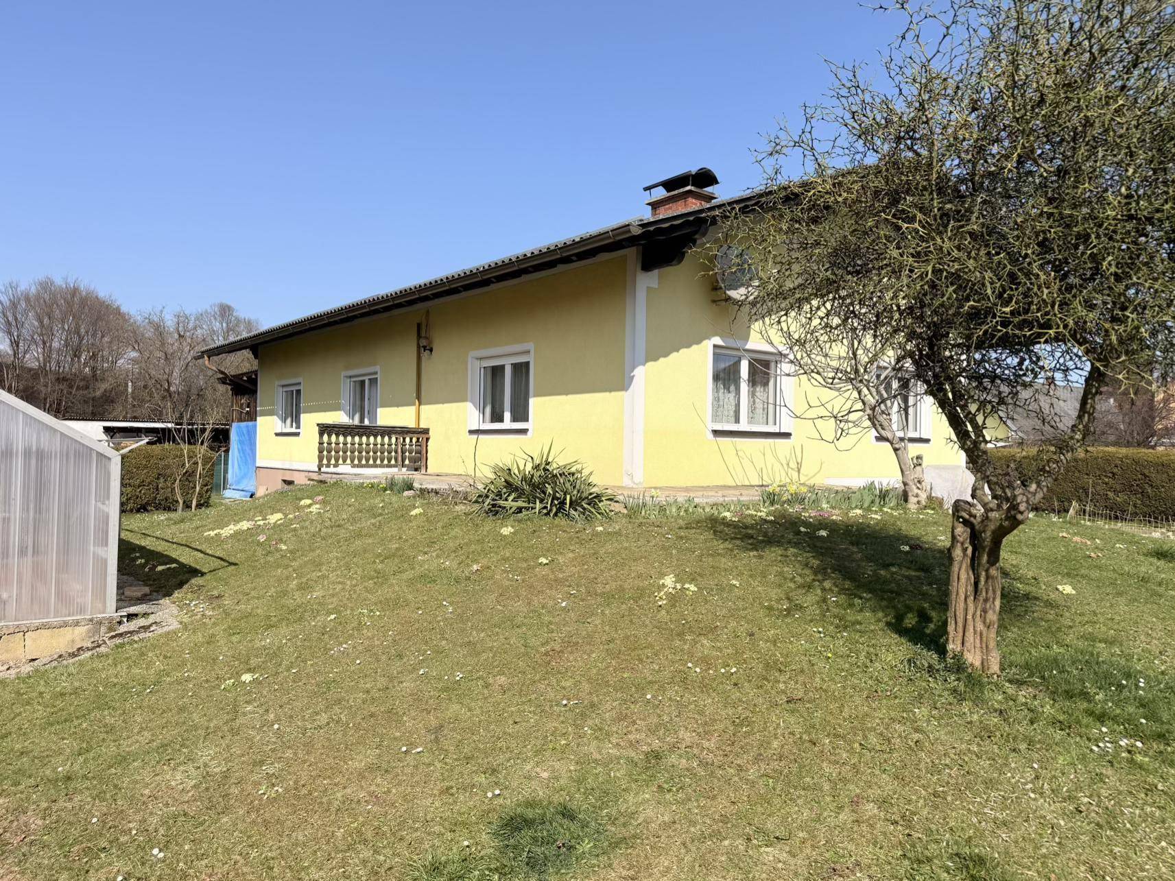 Seitenansicht des gelben Hauses mit einem grünen Rasenhügel und einem Baum im Vordergrund bei Sonnenschein.