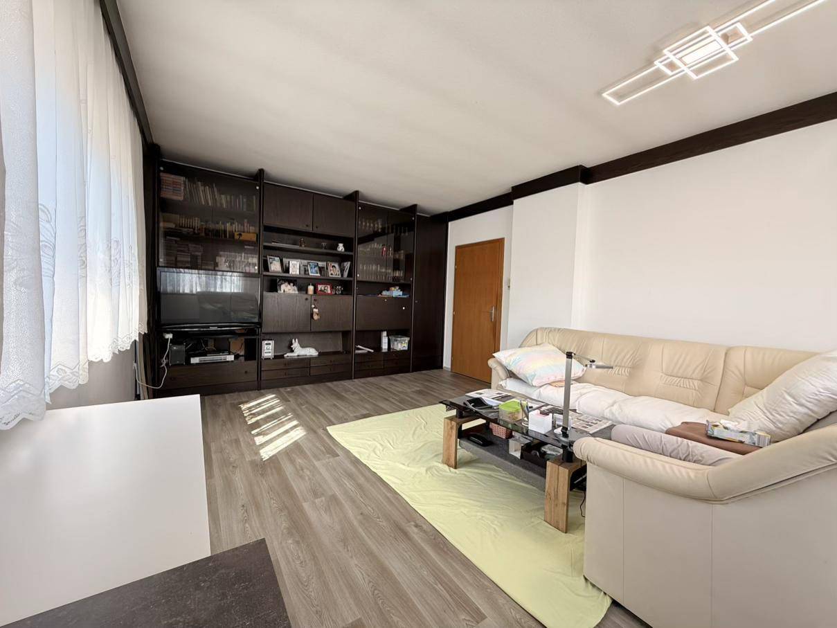 Wohnzimmer mit beigem Sofa, großer Schrankwand und Laminatboden, hell durch Tageslicht.