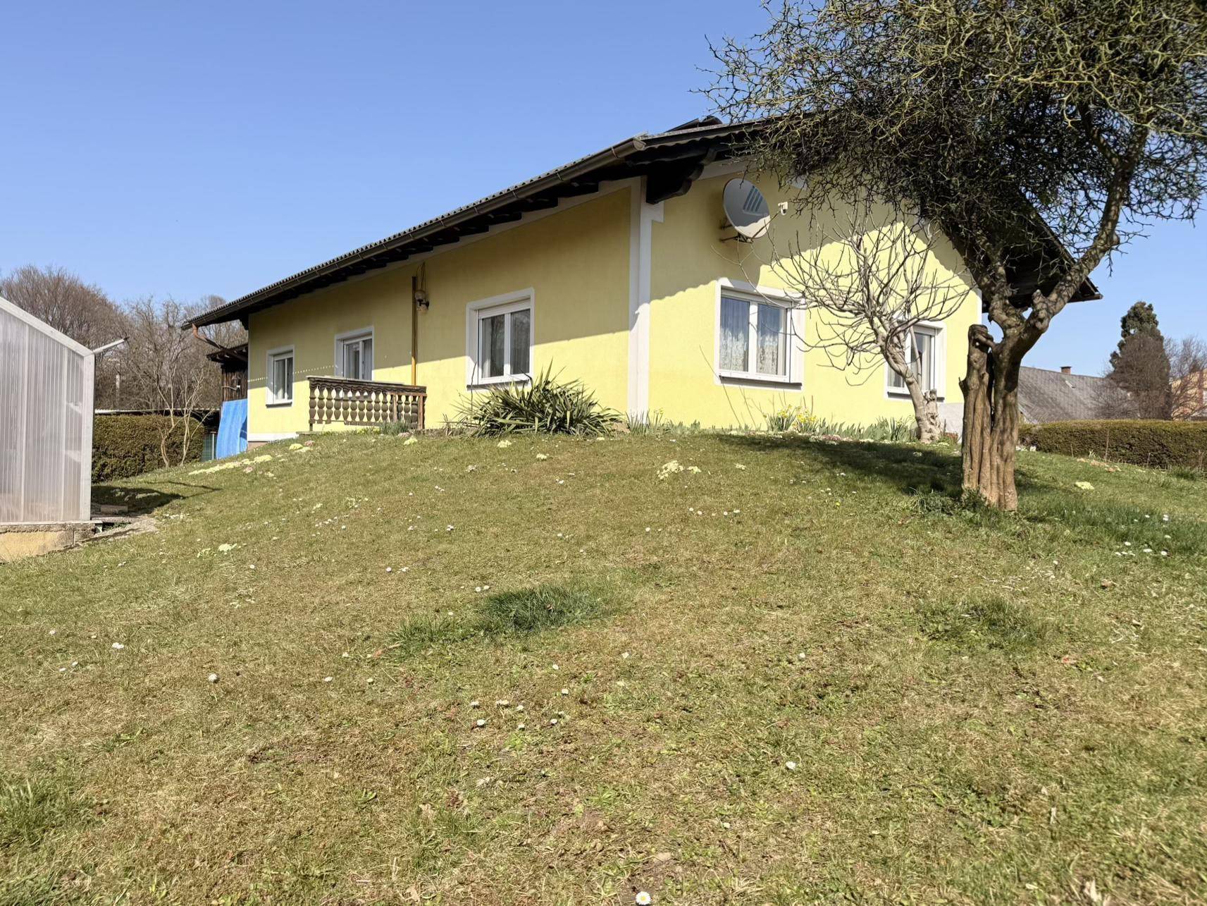 Seitenansicht des gelben Hauses mit einem grünen Rasenhügel und einem Baum im Vordergrund bei Sonnenschein.