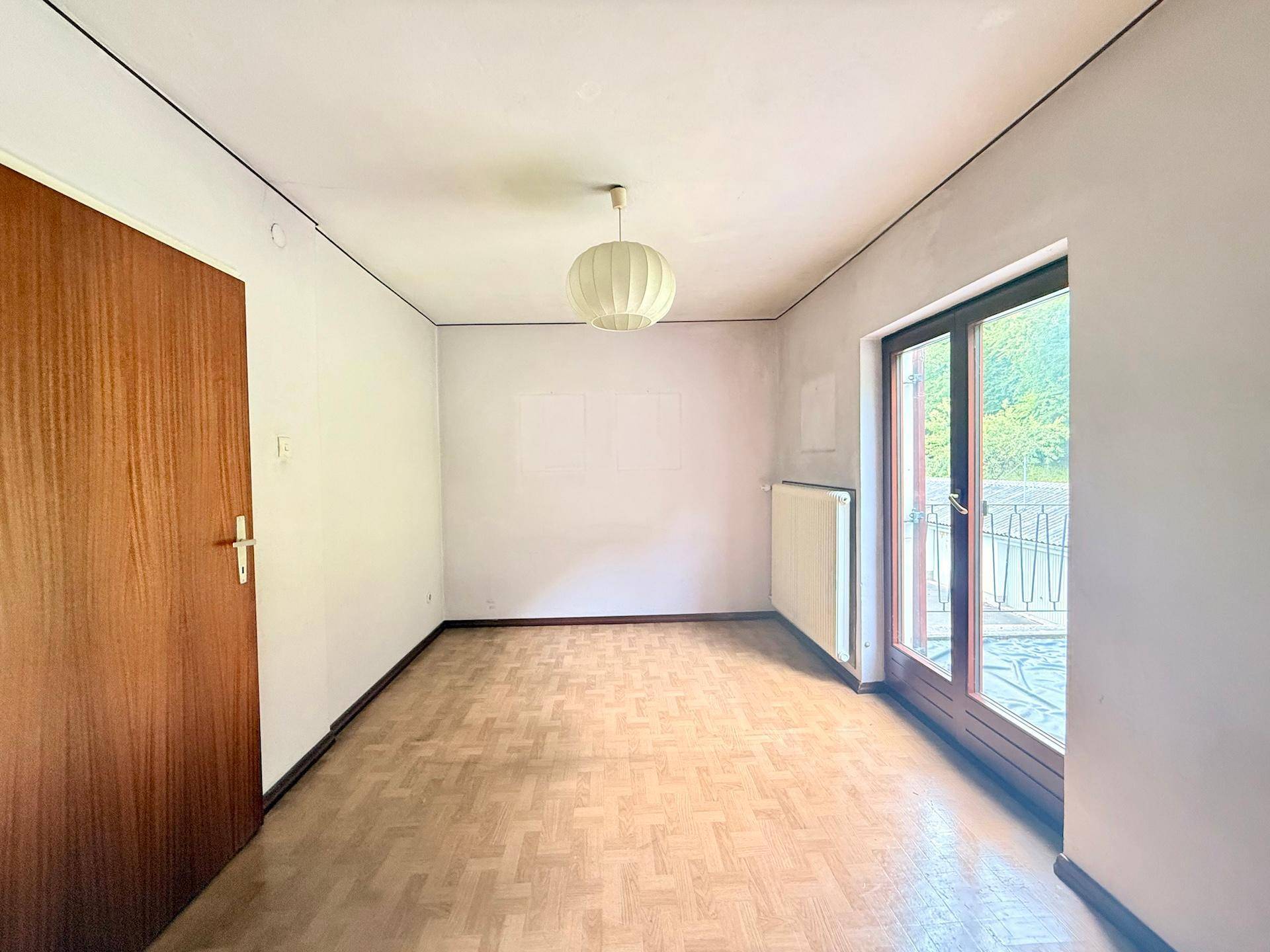 Helles Zimmer mit Parkettboden, Heizkörper und Zugang zu einem kleinen Balkon mit Blick ins Grüne.