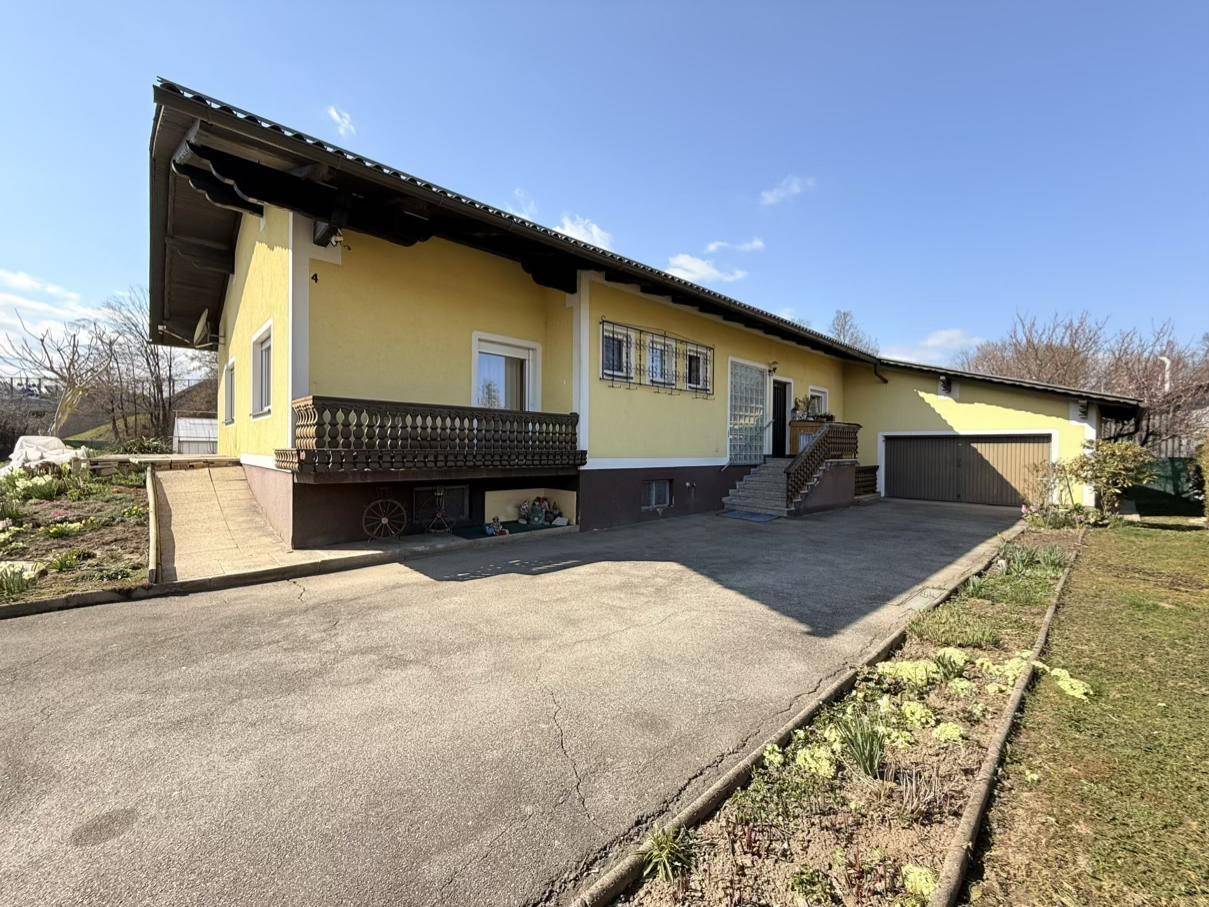 Ansicht des gelben Hauses mit der Einfahrt und einem kleinen Balkon mit Holzgeländer an einem sonnigen Tag.