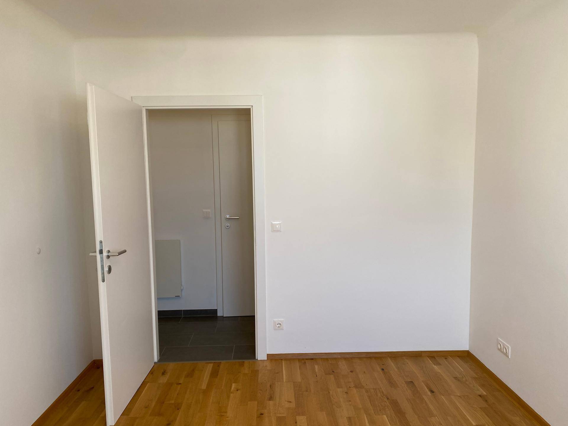 Helles Zimmer mit Parkettboden und weiß gestrichenen Wänden, mit Zugang zum Flur.