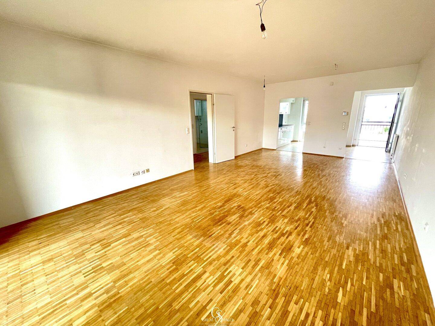 Geräumiges Wohnzimmer mit hellem Parkettboden und Zugang zu weiteren Räumen und dem Balkon.
