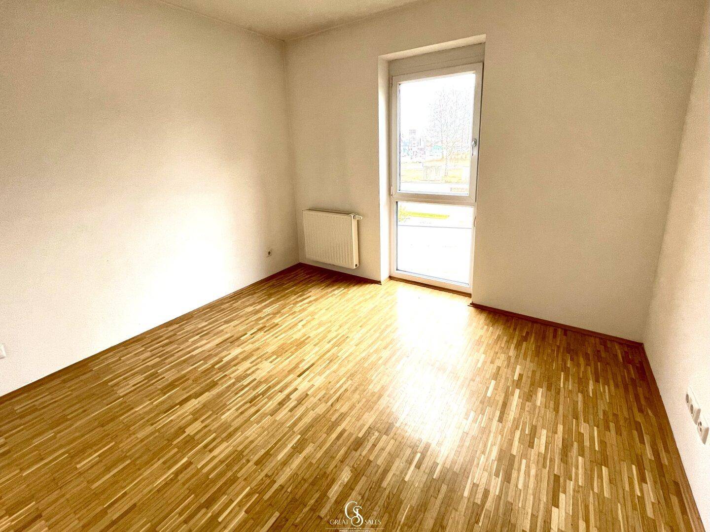 Helles Schlafzimmer mit Parkettboden und großem Fenster, das viel Tageslicht hereinlässt.