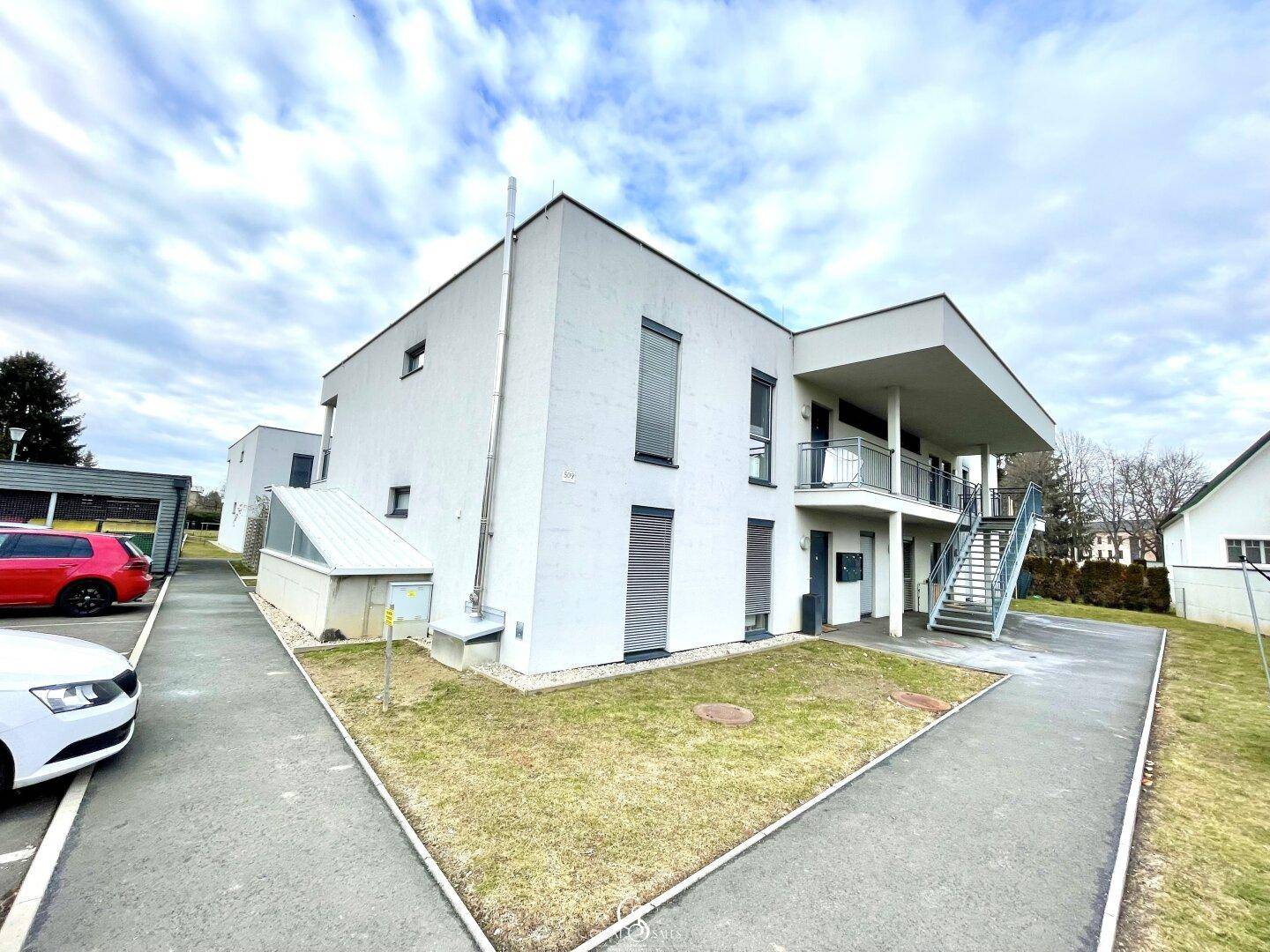 Modernes Mehrfamilienhaus mit weißer Fassade und Außenbereich, umgeben von Grünflächen.