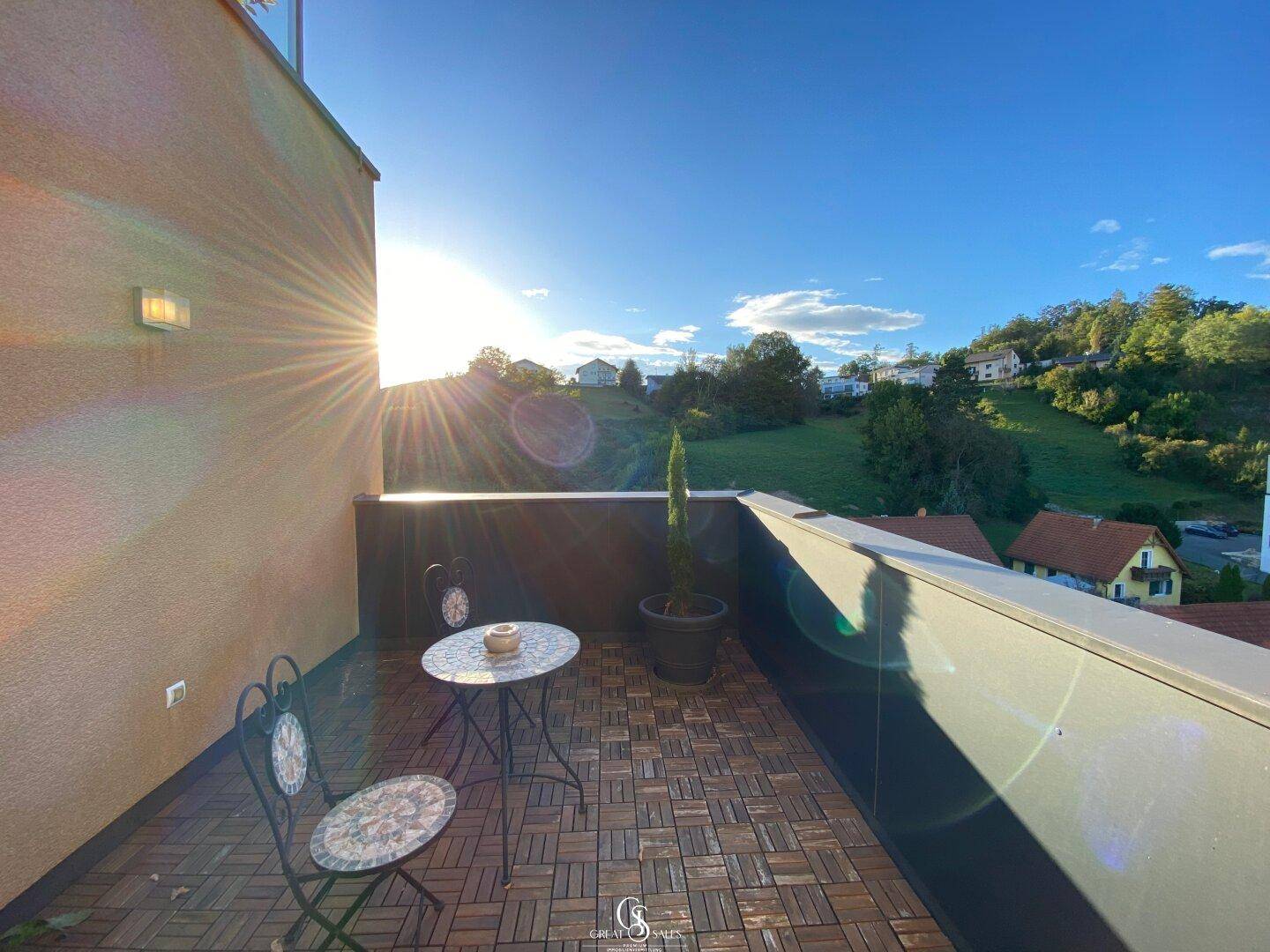 Sonniger Balkon mit Holzboden, kleinem Tisch und Stühlen, umgeben von grüner Landschaft.