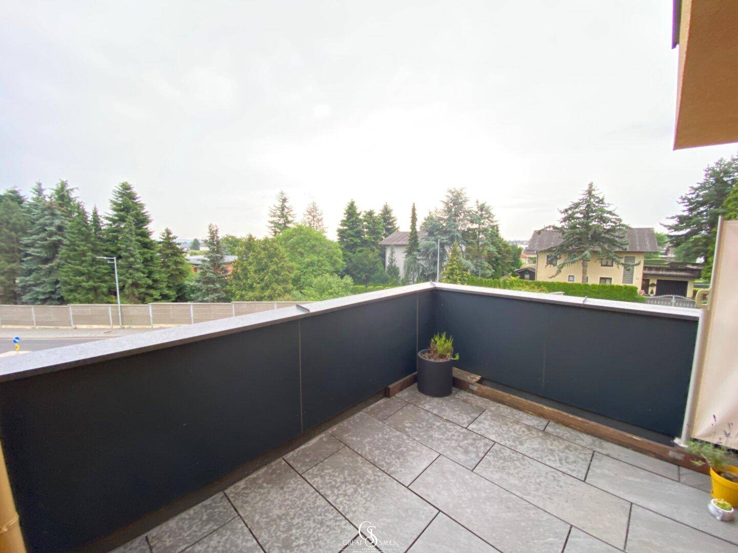 Balkon mit Fliesenboden und einer kleinen Pflanze, umgeben von Bäumen und Häusern.