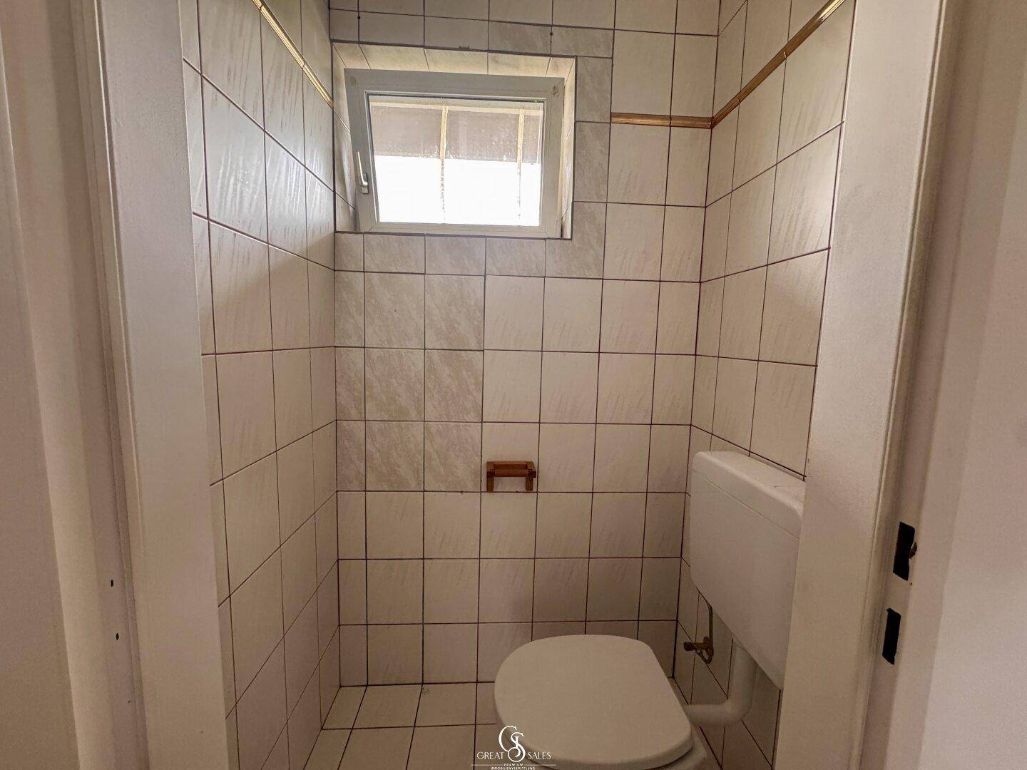 Kleines WC mit Fenster, weißen Wandfliesen und einer einfachen Toilette.
