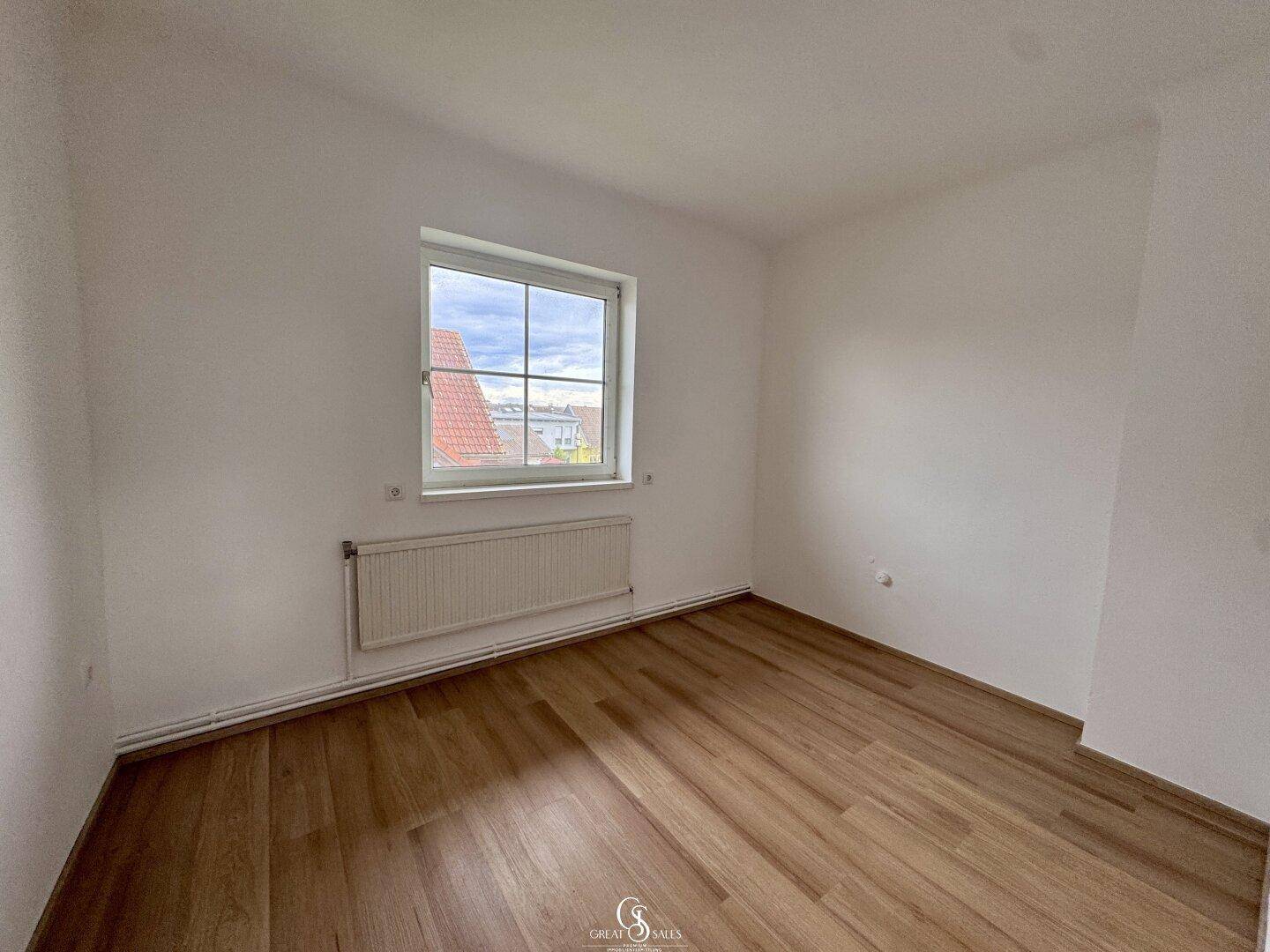 Helles Zimmer mit Laminatboden und Fenster, das einen Blick ins Grüne bietet.