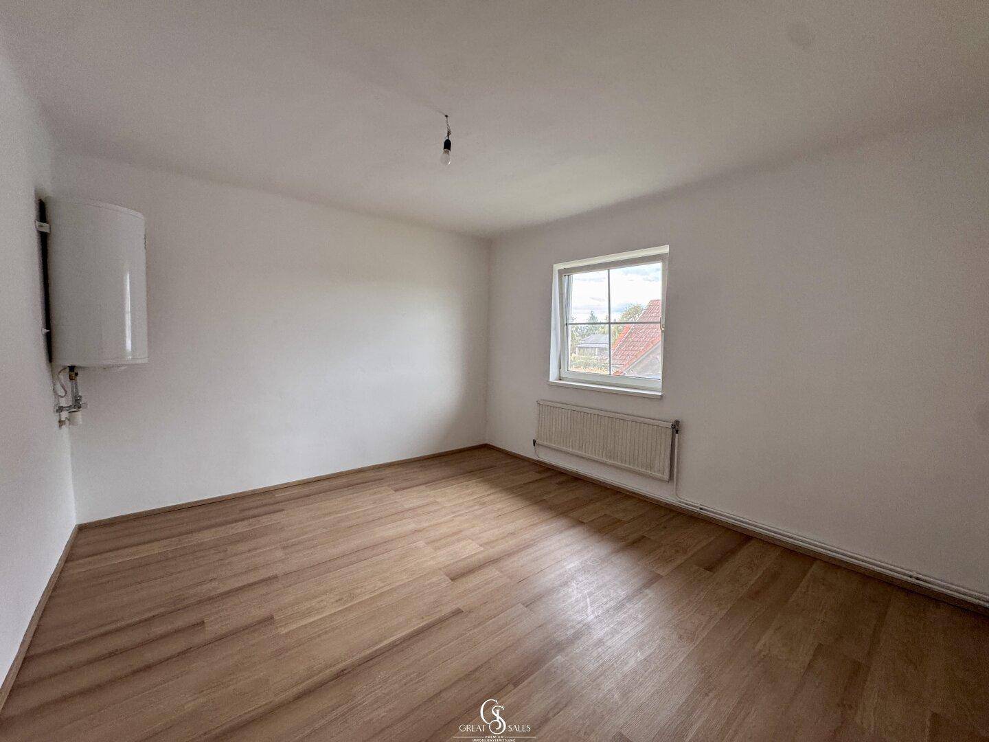 Helles Zimmer mit Laminatboden, Fenster und einem Warmwasserboiler an der Wand.