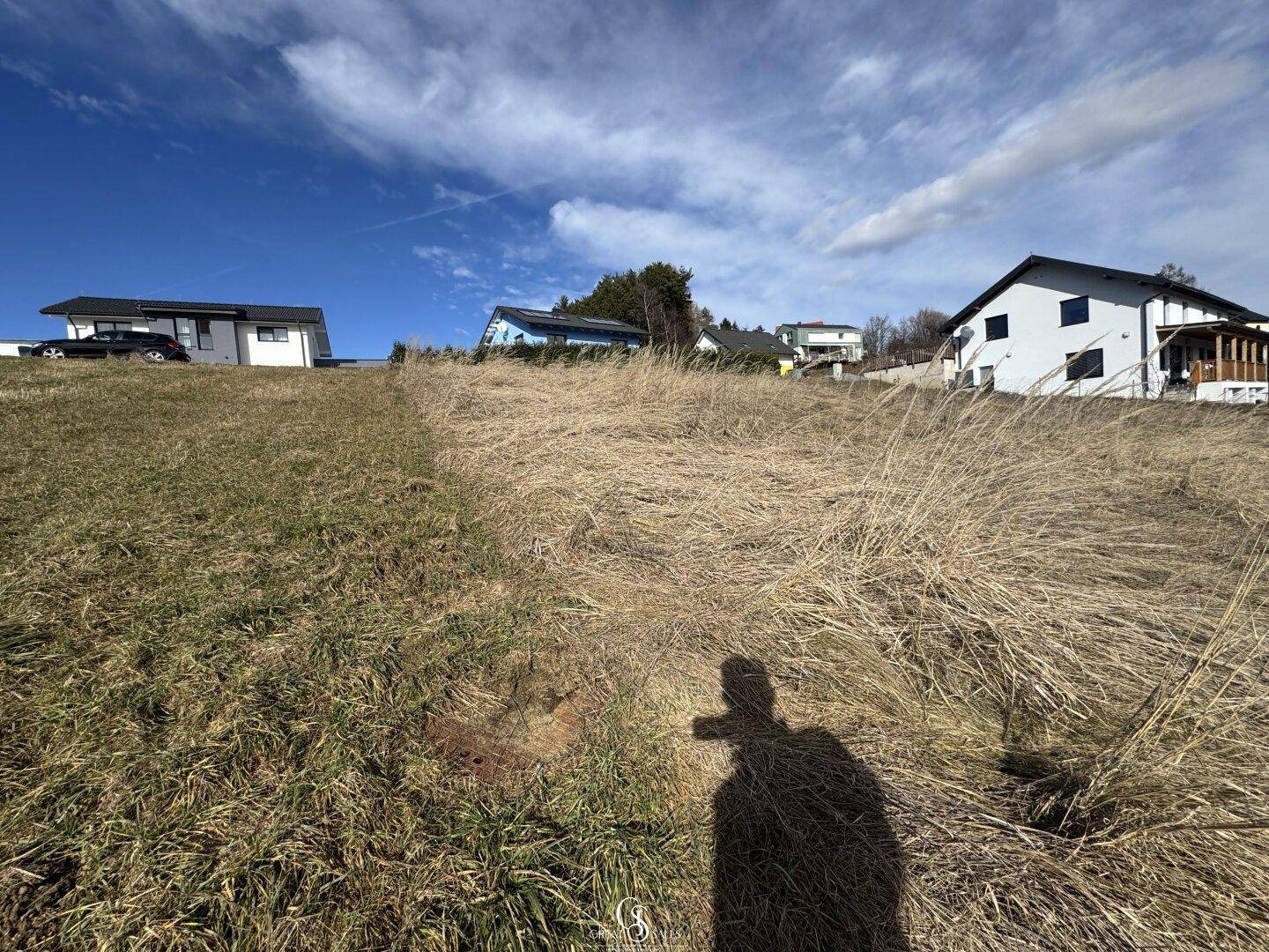 Sanfte Hügellandschaft mit trockenem Gras und mehreren Wohnhäusern am Horizont unter blauem Himmel.