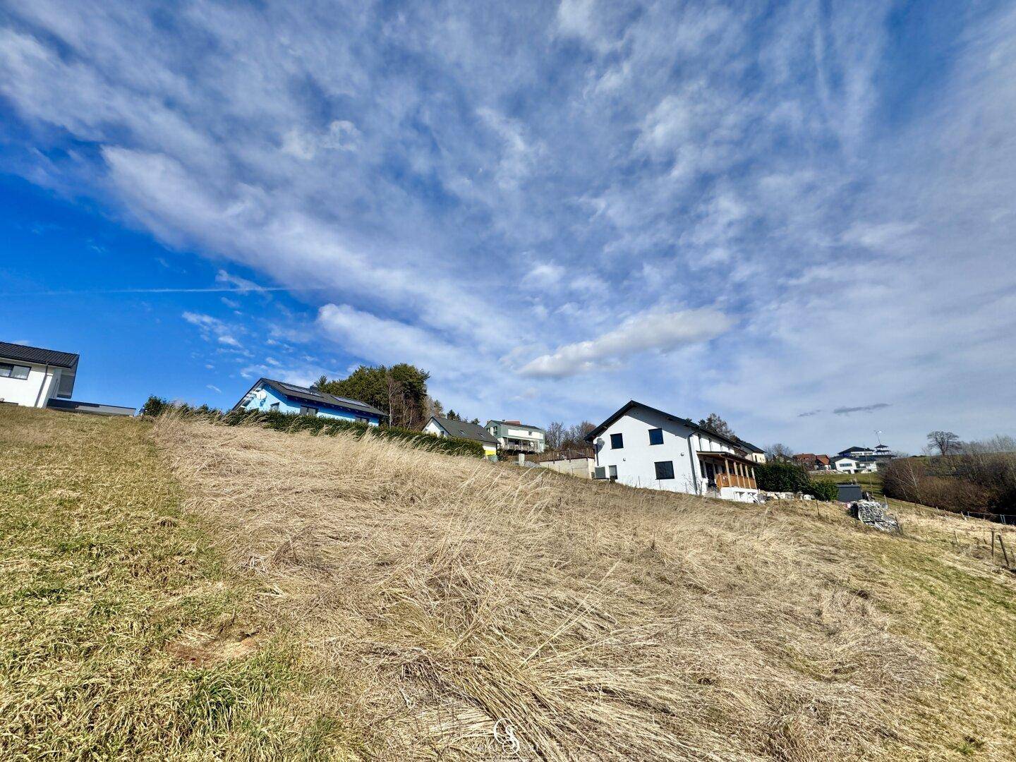 Weitläufige Hügellandschaft mit mehreren Wohnhäusern am Horizont unter einem blauen Himmel.