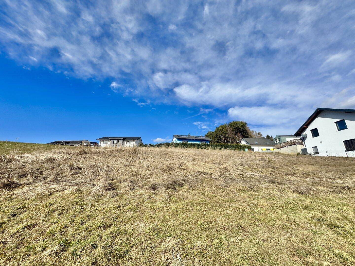 Sanfte Hügel mit trockenem Gras und einer Reihe von Häusern in der Ferne unter weitem Himmel.