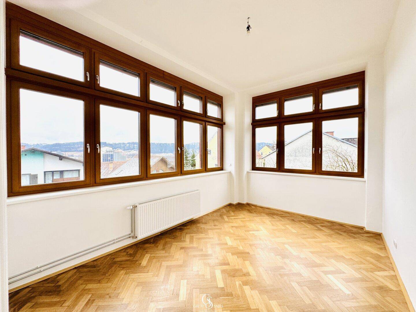 Heller Raum mit Fischgrätparkett und großen Holzfenstern, die viel Tageslicht hereinlassen.