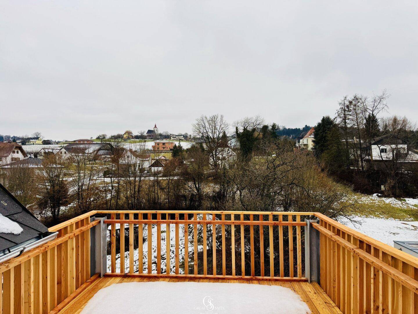 Großer Holzbalkon mit weitem Blick über die verschneite Landschaft und ein Dorf in der Ferne.