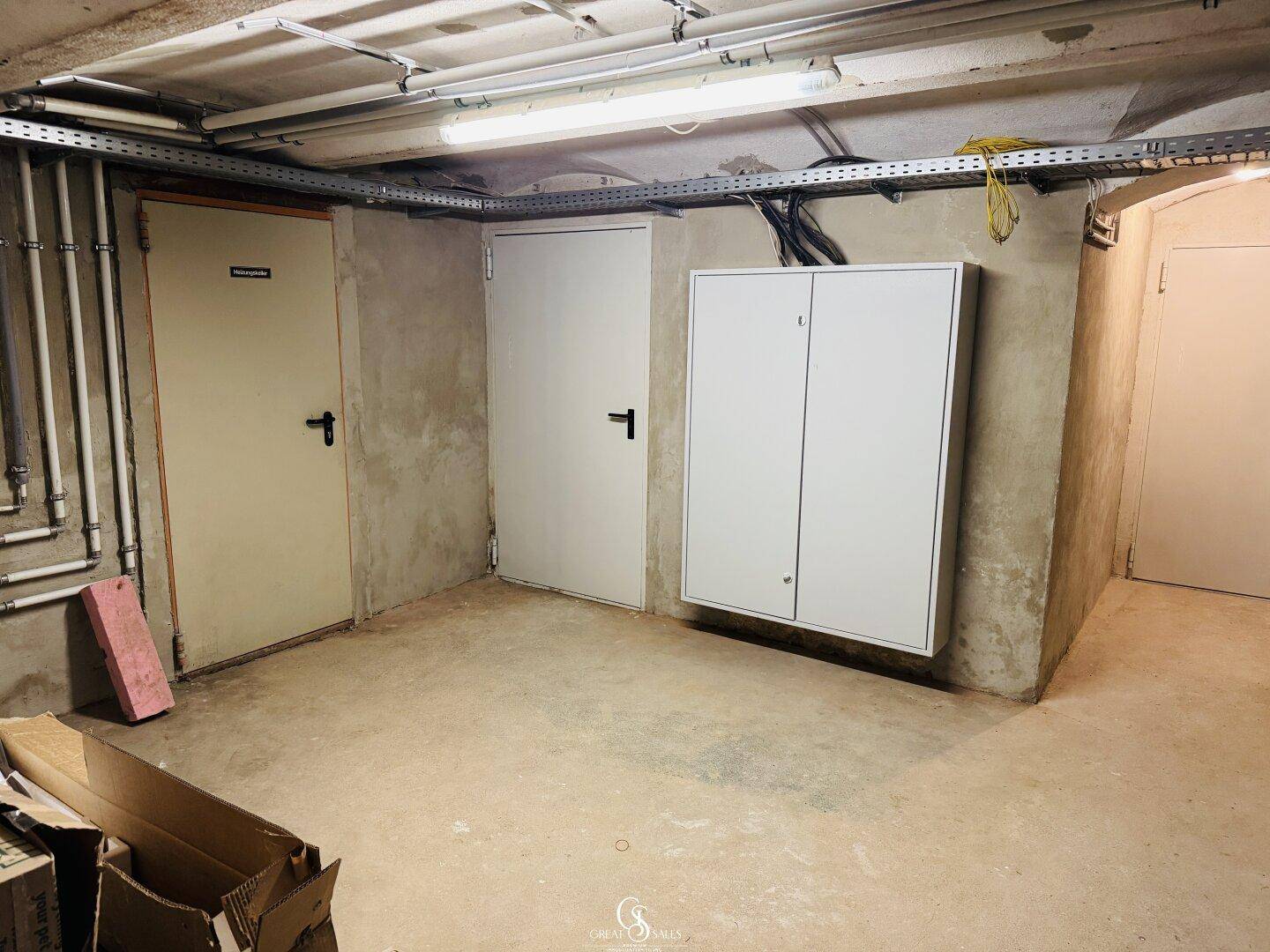 Kellerraum mit Betonboden und sichtbaren Leitungen, dient als Technik- oder Abstellraum.