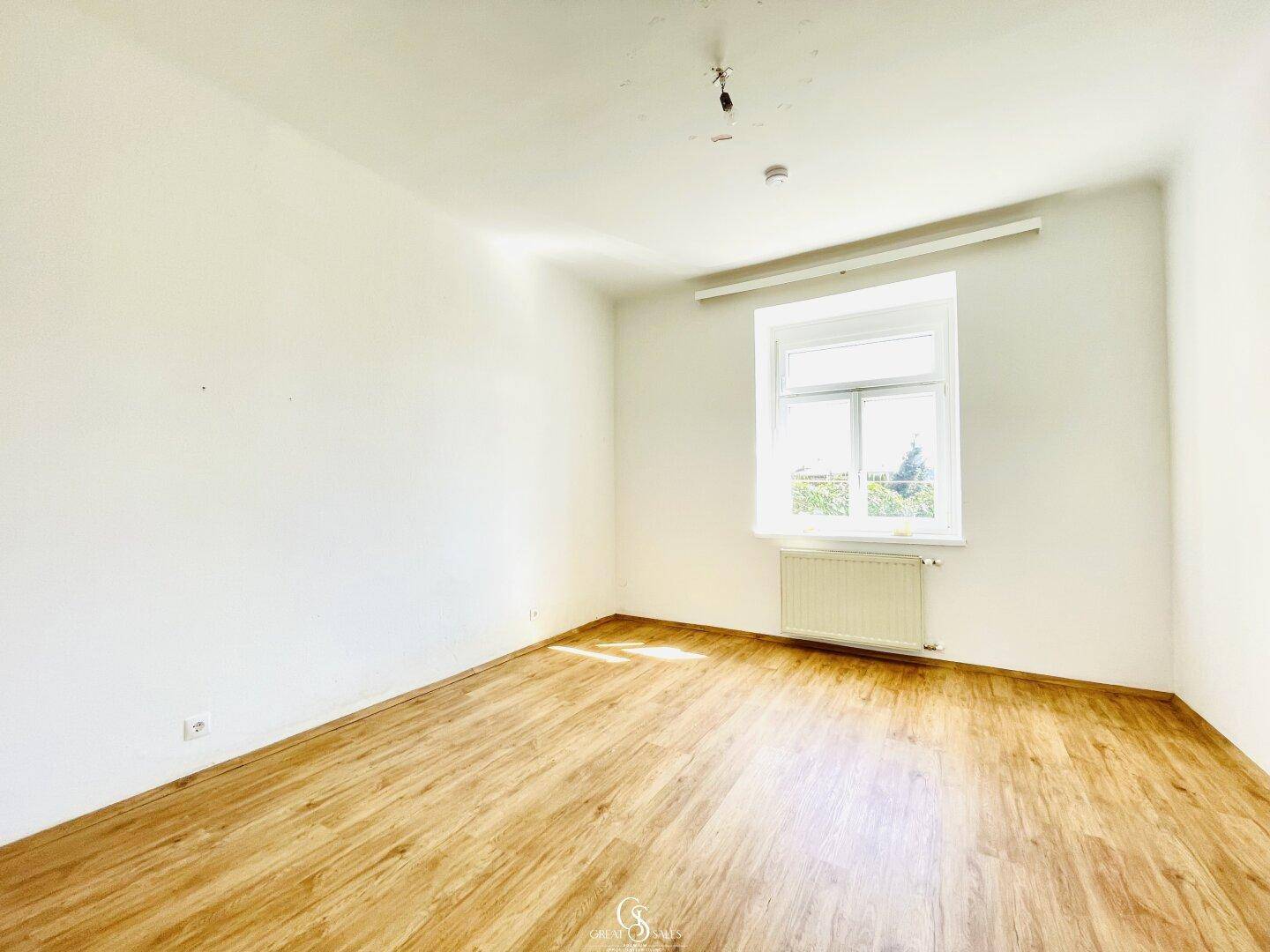 Geräumiges, helles Zimmer mit Holzparkettboden und einem großen Fenster, das viel Licht hereinlässt.