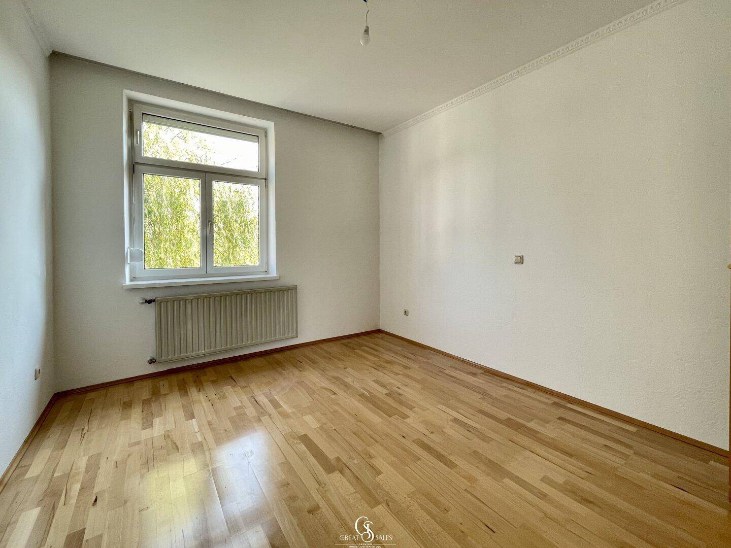 Geräumiges Schlafzimmer mit Holzparkettboden, weiß gestrichenen Wänden und einem Fenster mit Blick ins Grüne.