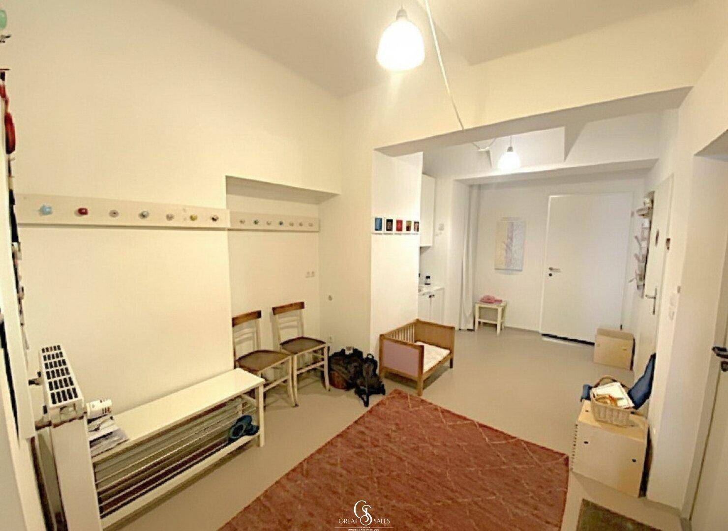 Heller Mehrzweckraum mit Garderobenhaken, Sitzbank und einer kleinen Küchenzeile im Hintergrund.