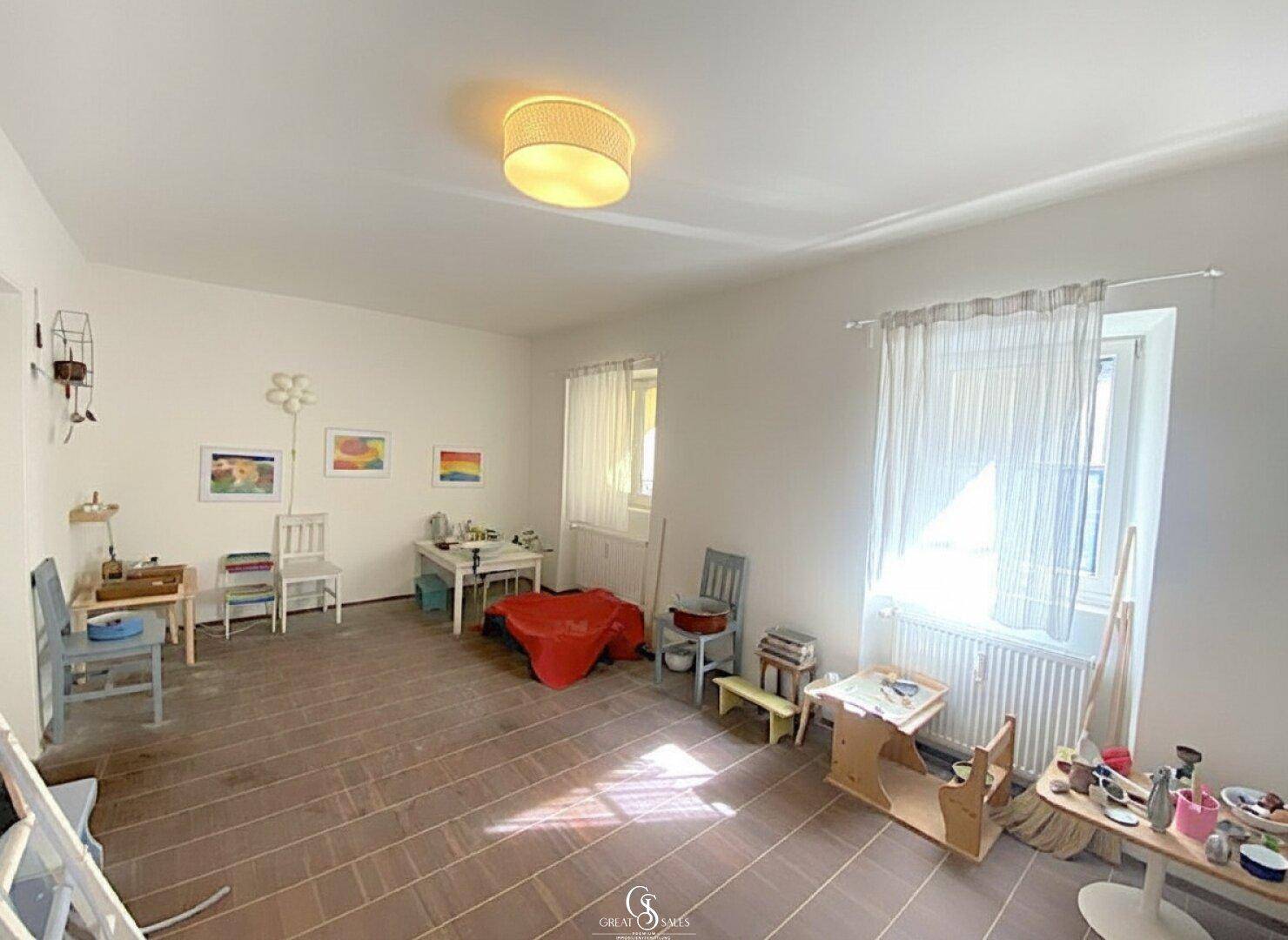 Geräumiger Spielraum mit viel Tageslicht, Kindermöbeln und dunklem Fliesenboden.
