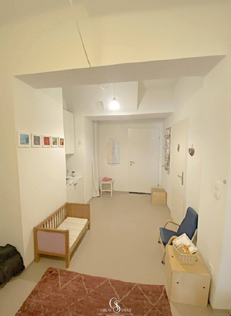 Heller Raum mit Kinderbett, Stuhl und einer kleinen Küchenzeile, gut beleuchtet und funktional.