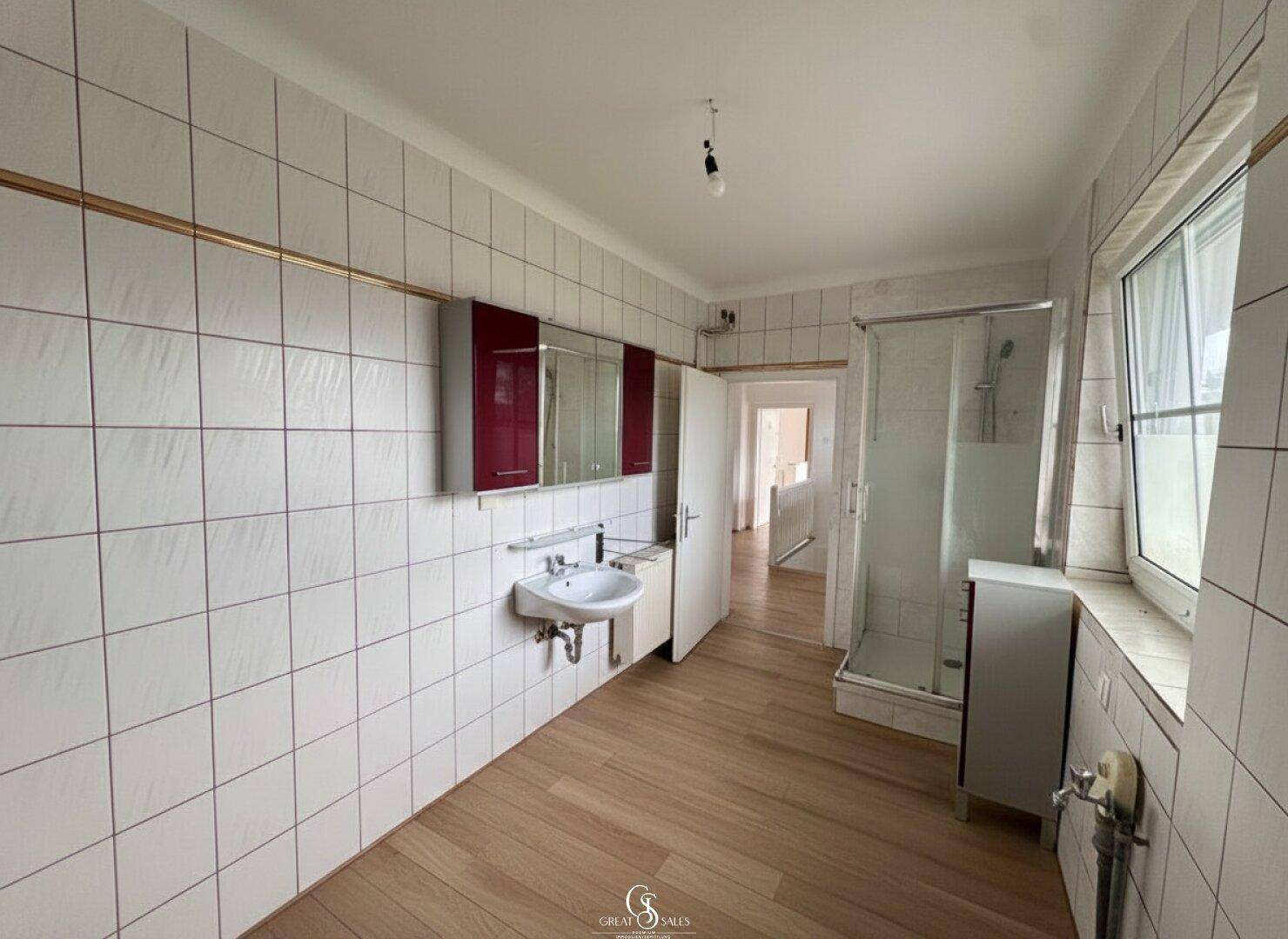 Helles Badezimmer mit weißen Fliesen, Holzfußboden, Duschkabine und Fenster für Tageslicht.