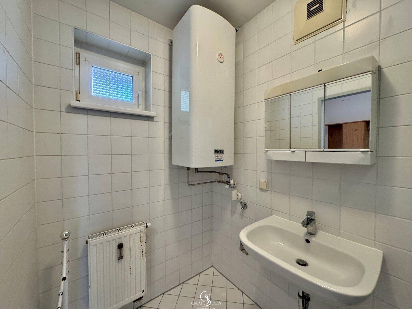 Badezimmer mit Fenster, Waschbecken und Boiler, komplett gefliest für einfache Pflege.