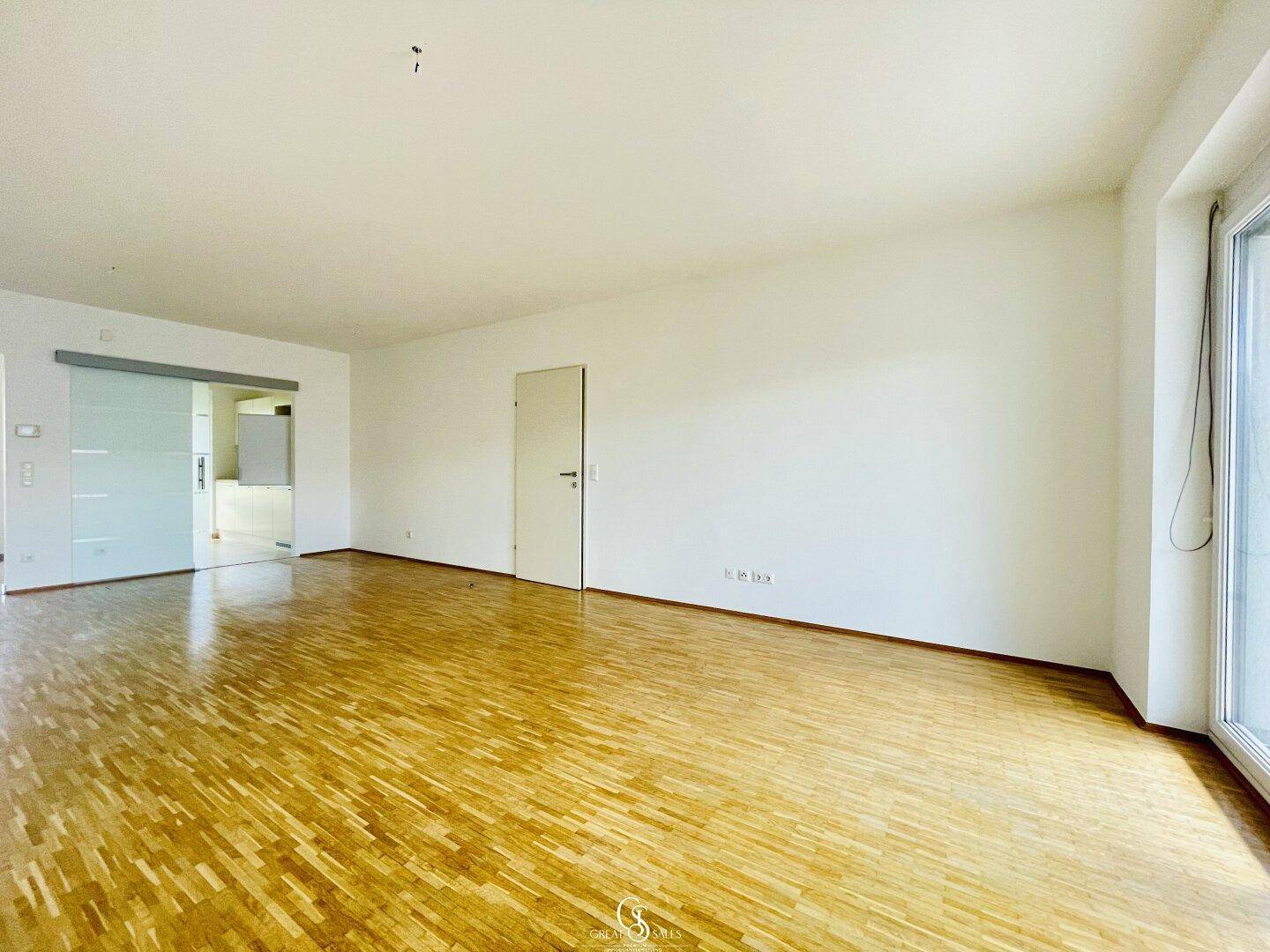 Lichtdurchflutetes Wohnzimmer mit Parkettboden und Glasschiebetür zur angrenzenden Küche.