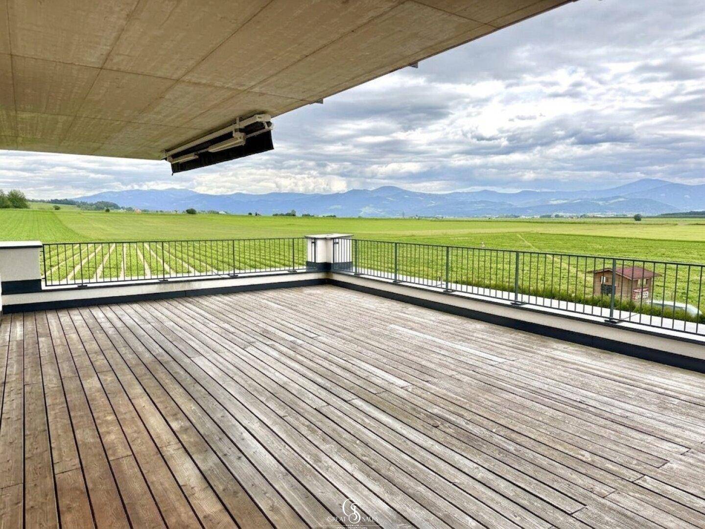 Weitläufige Holzterrasse mit Panoramablick über grüne Felder und die majestätischen Berge.
